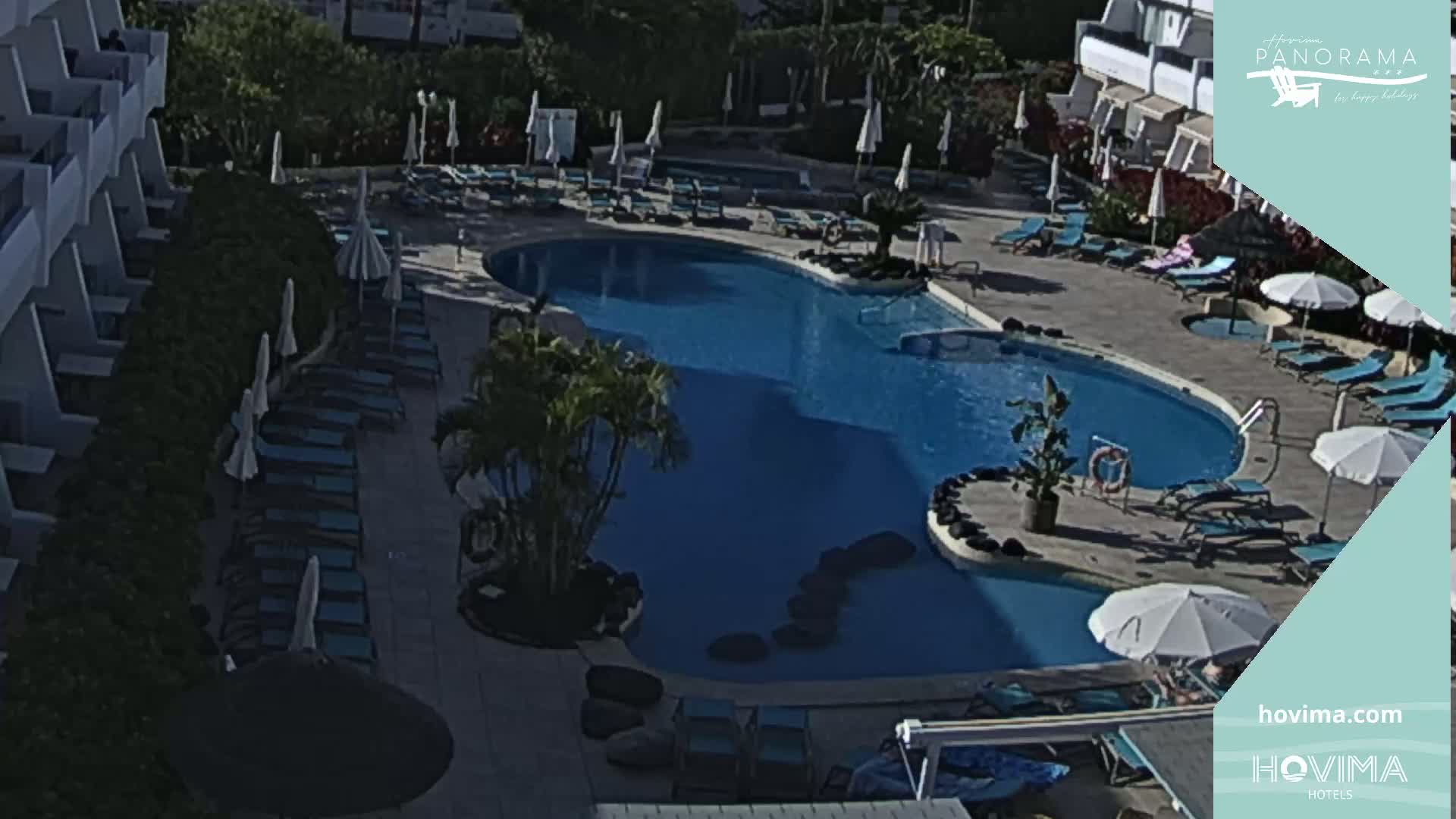An outdoor swimming pool surrounded by lounge chairs and umbrellas, next to a building, on a sunny day.