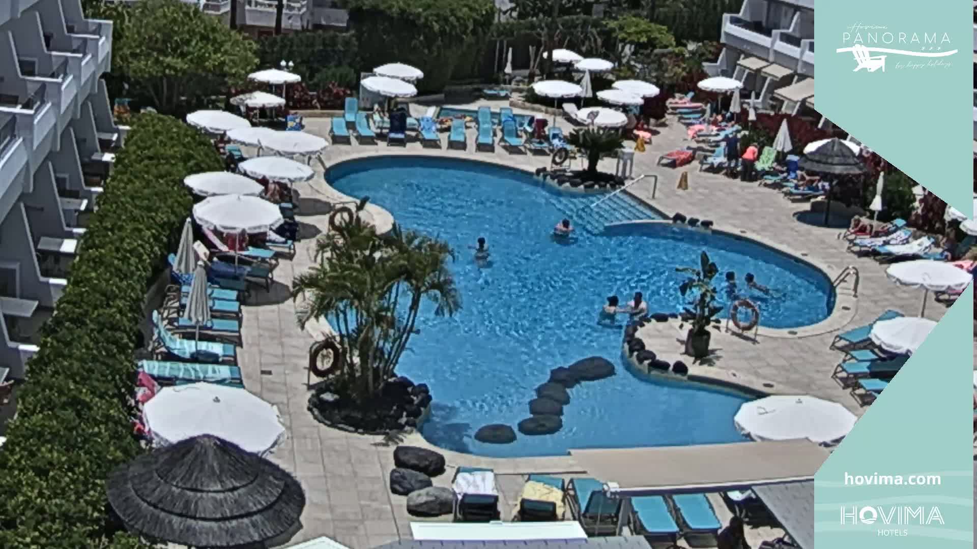 An outdoor pool with many people swimming and lounging under umbrellas on a sunny day.