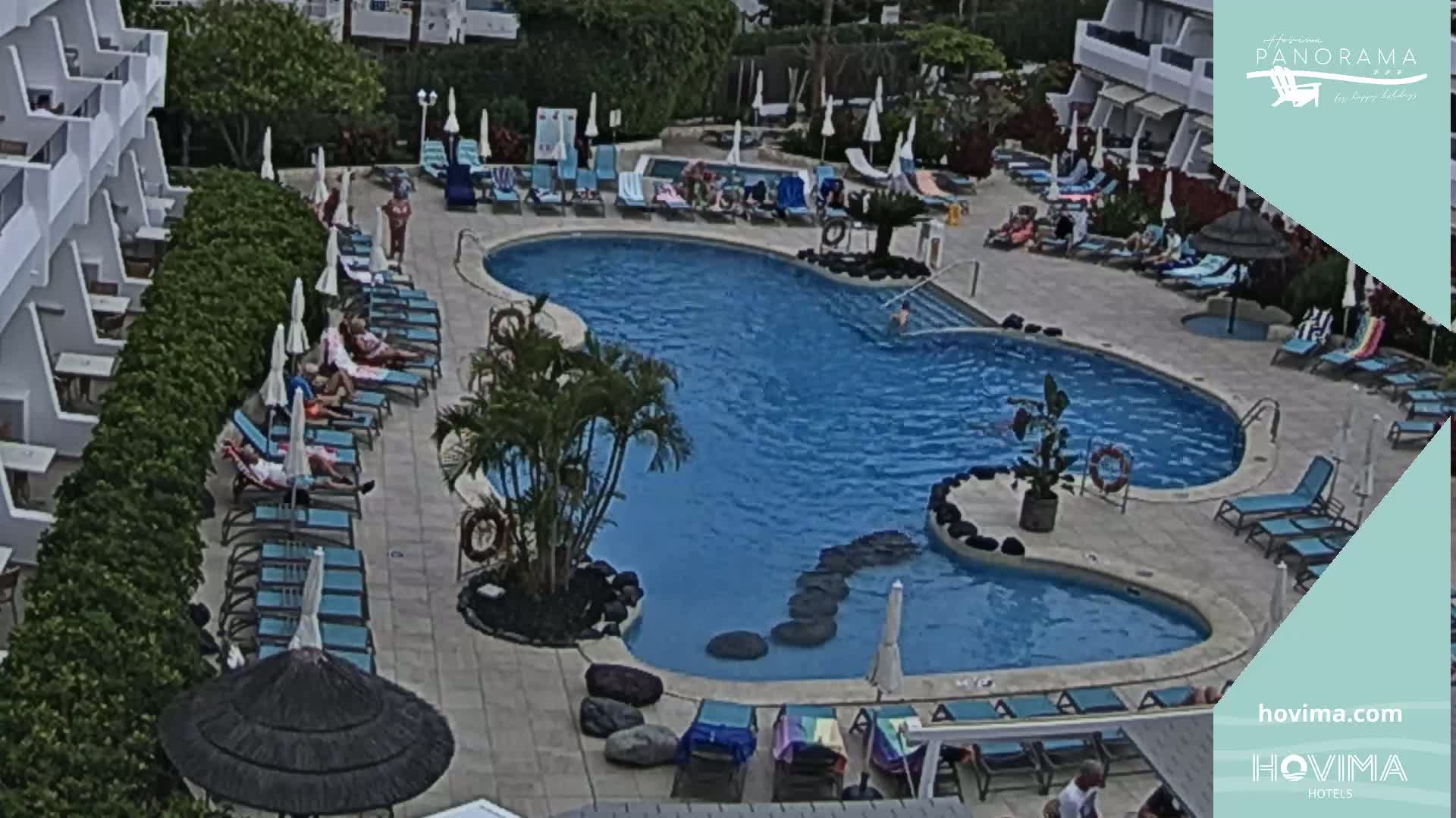 An outdoor swimming pool with surrounding lounge chairs and umbrellas, partially shaded by trees and buildings, on a sunny day.