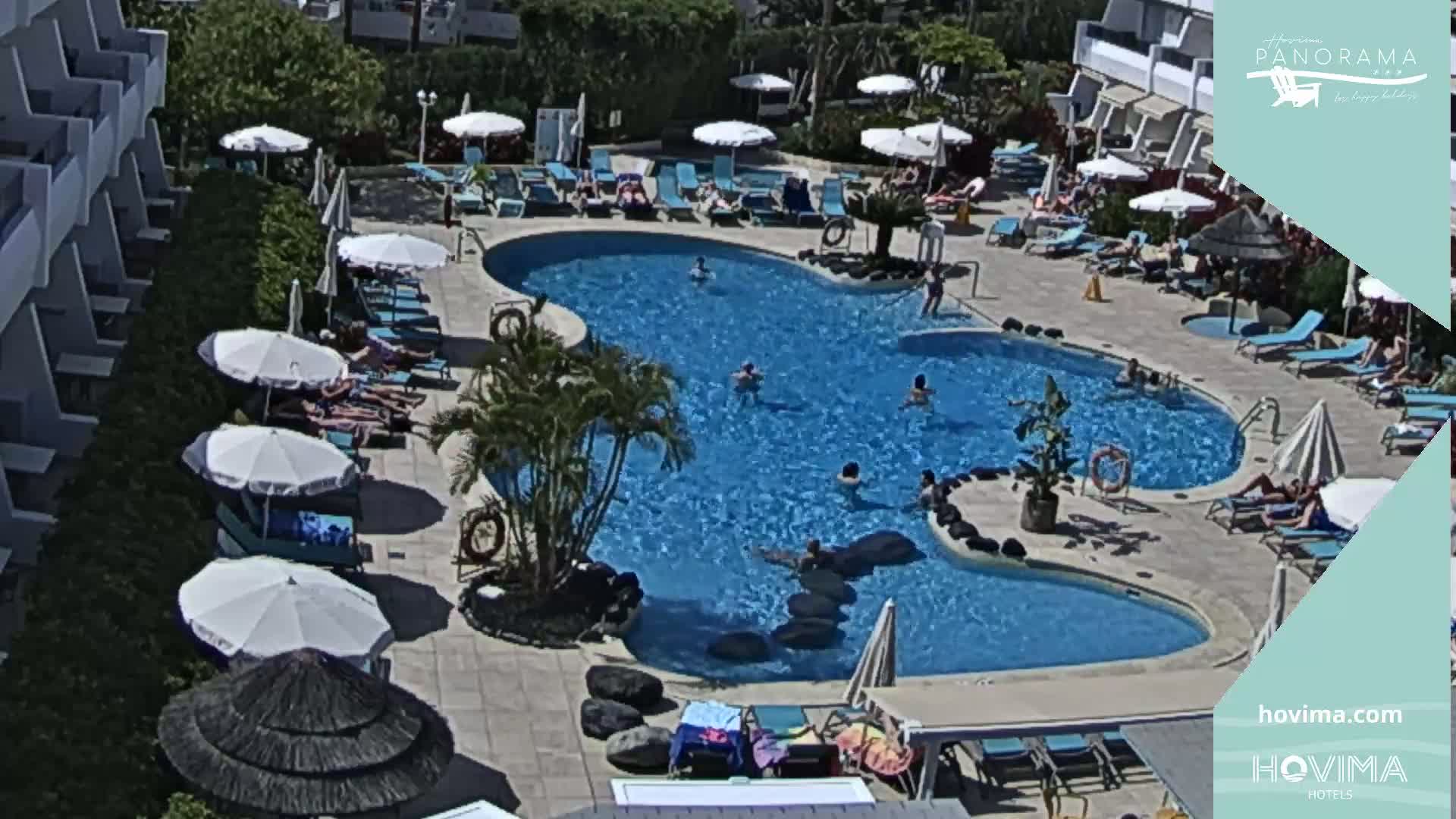 An outdoor swimming pool with several people swimming and sunbathing under umbrellas on a sunny day.