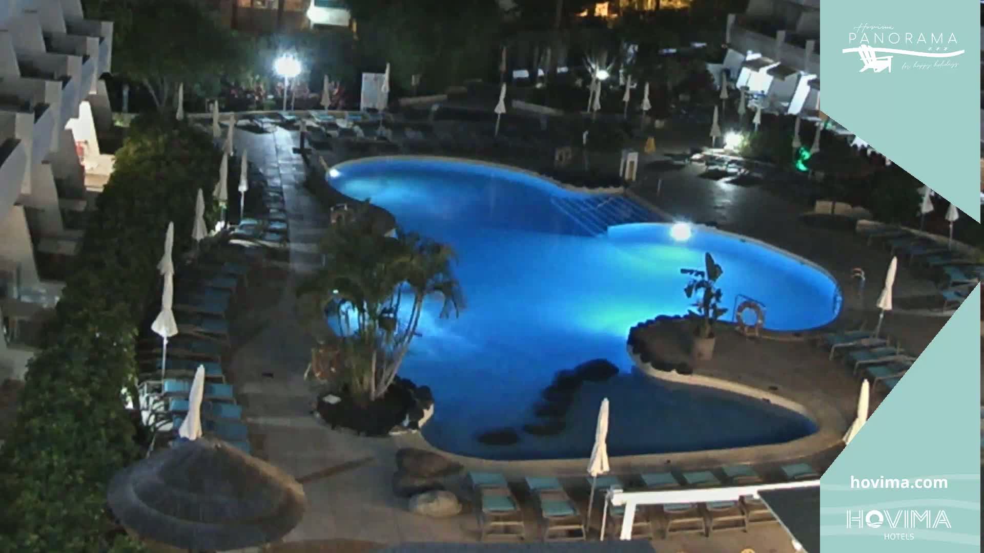 An illuminated, kidney-shaped swimming pool at night is surrounded by lounge chairs and umbrellas near a building.