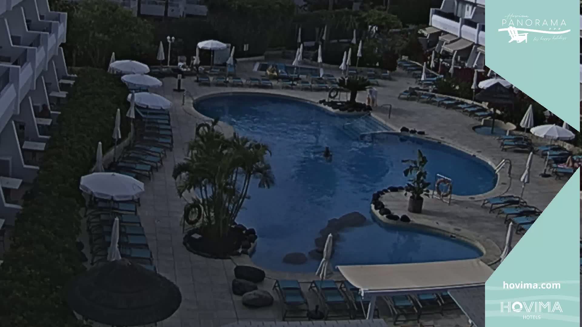 A resort pool area with lounge chairs, umbrellas, and palm trees, under a partly cloudy sky.