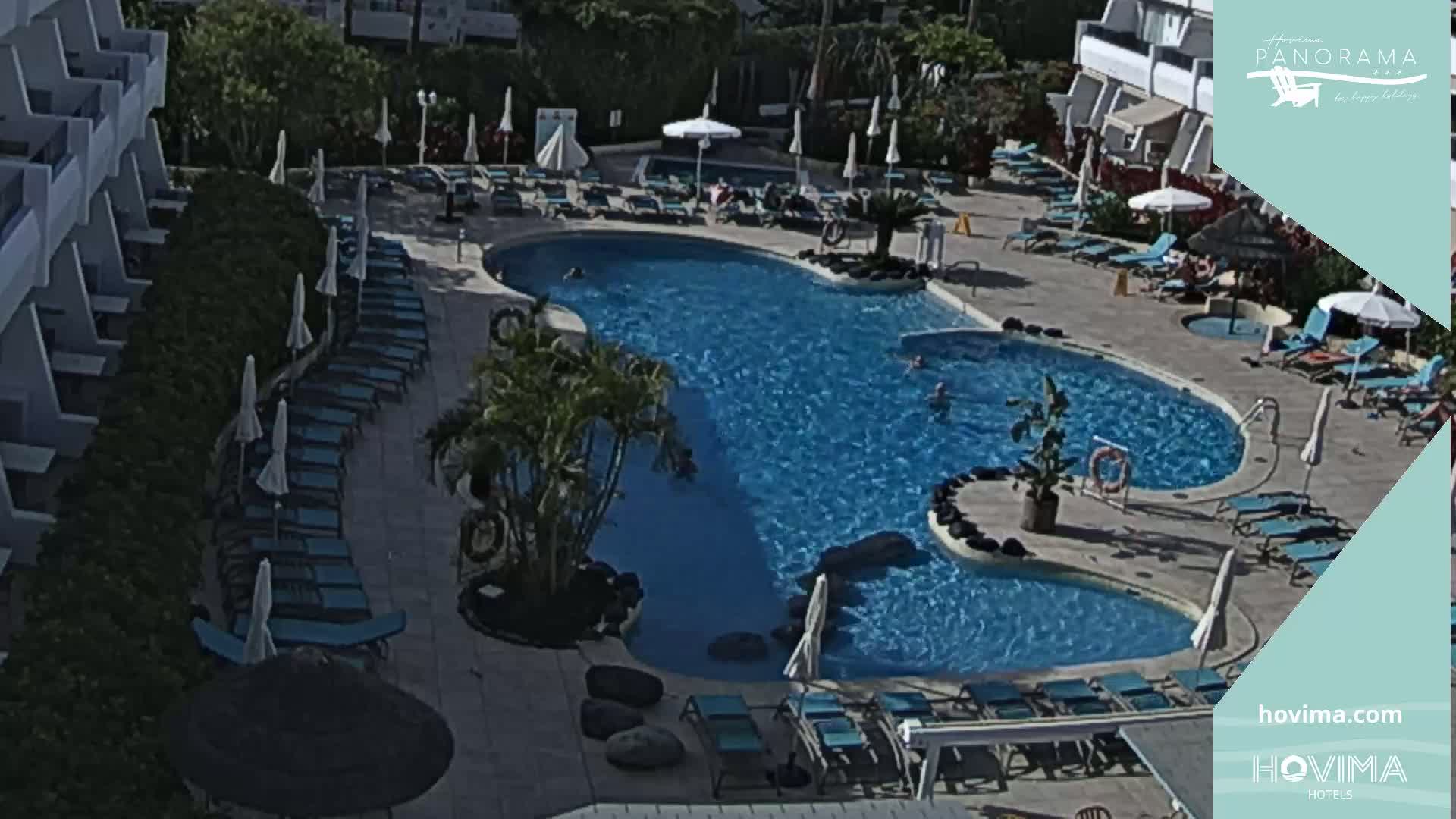 An outdoor swimming pool with surrounding lounge chairs and umbrellas is situated next to a multi-story building on a sunny day.