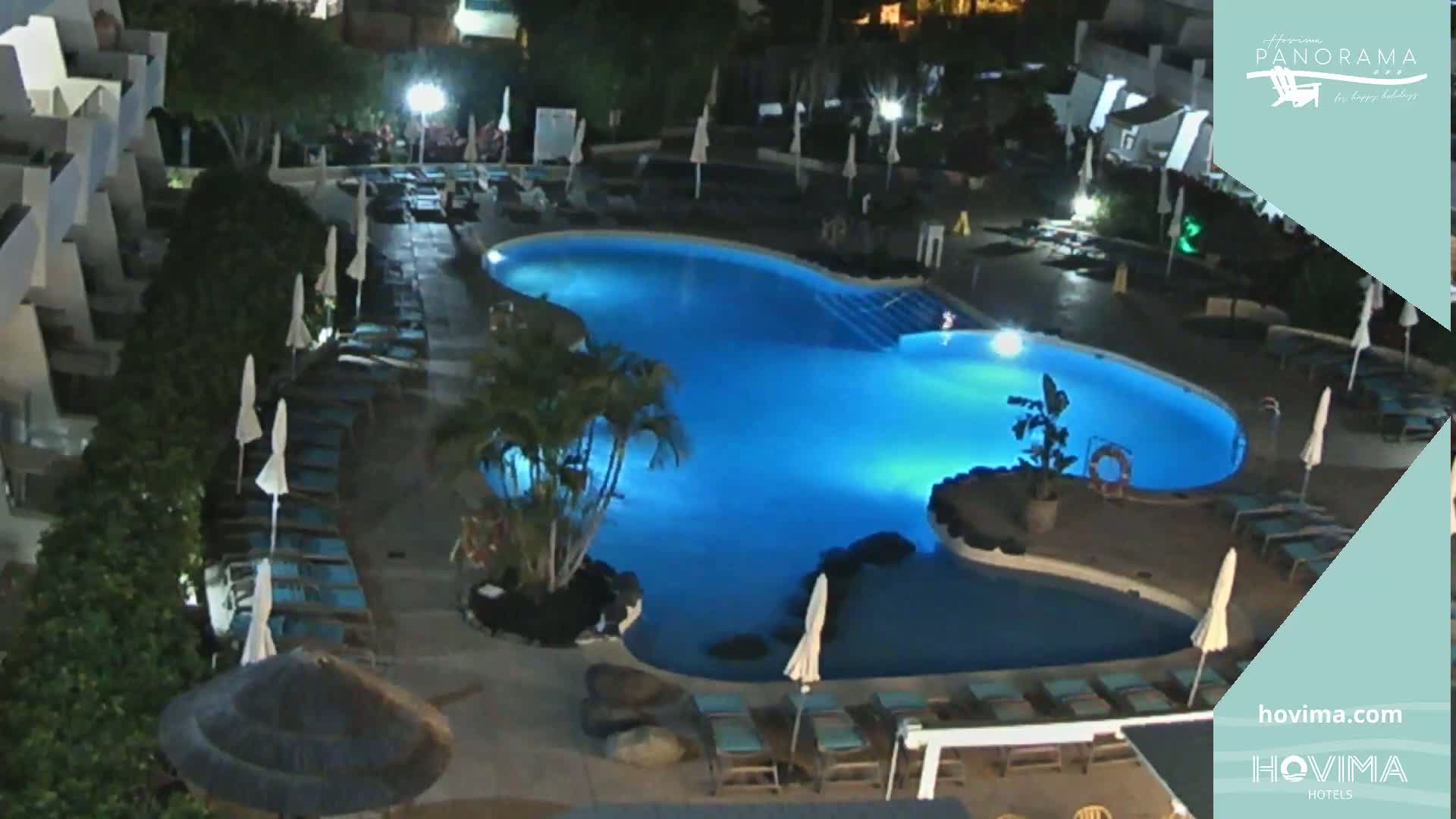 A nighttime view of an illuminated swimming pool surrounded by lounge chairs and palm trees at a resort.