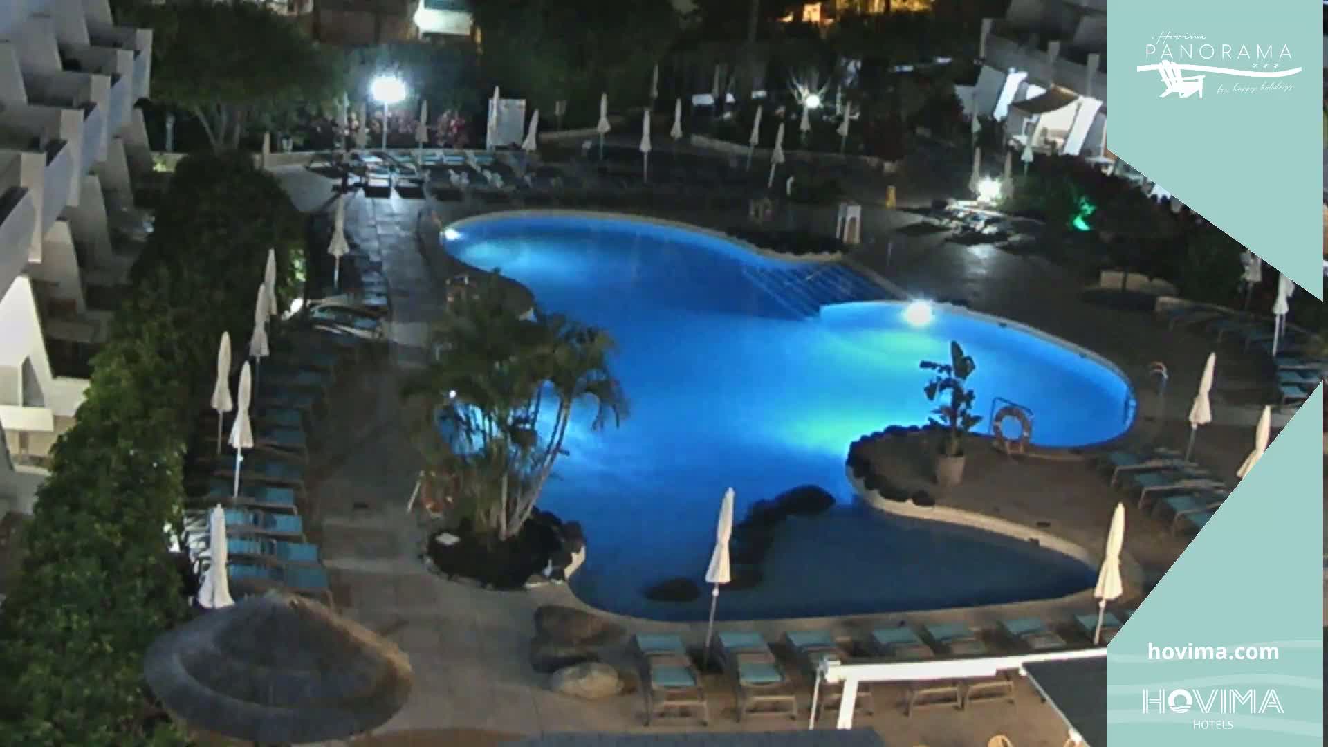An illuminated swimming pool surrounded by lounge chairs and umbrellas at night.