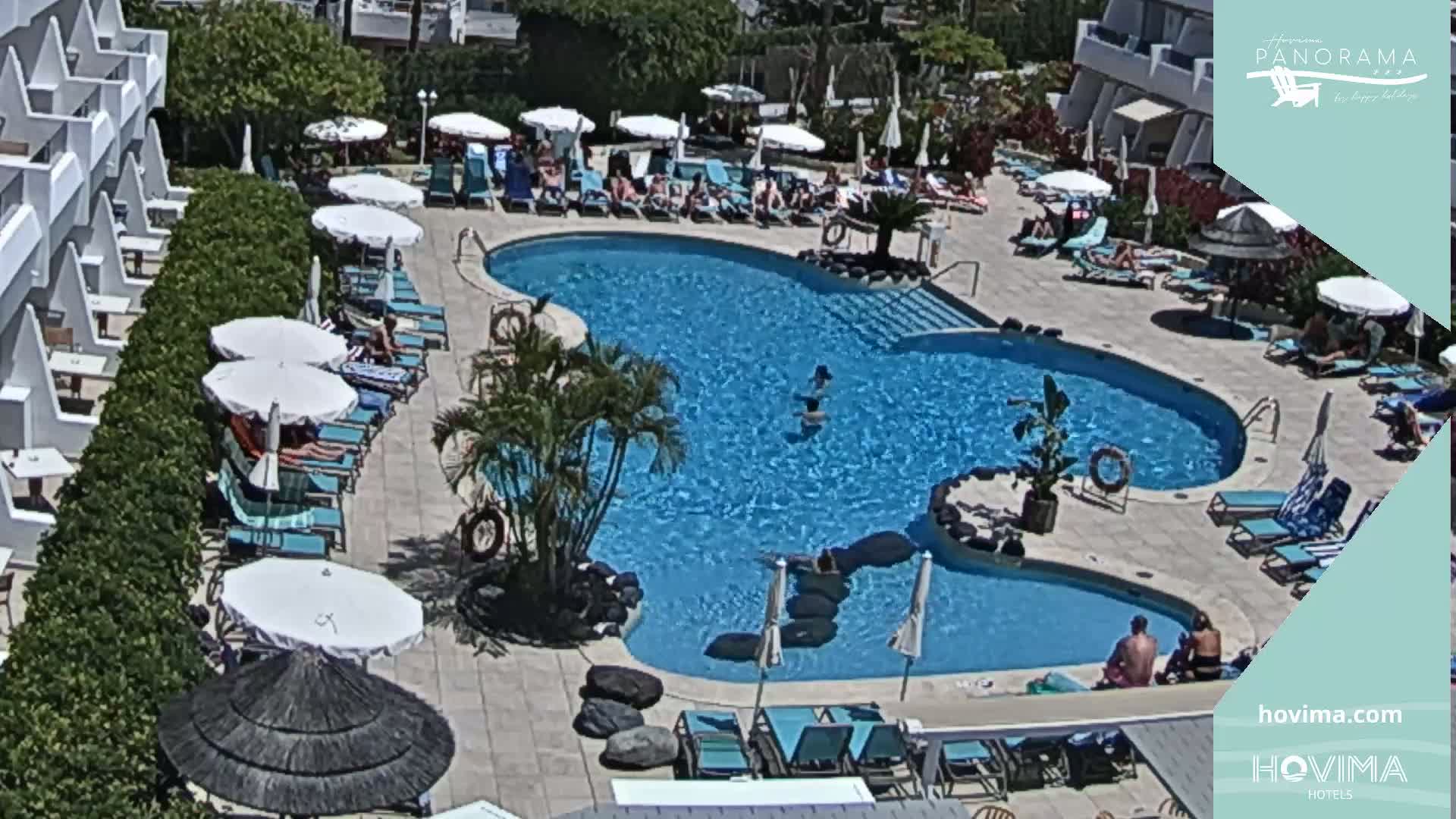 An overhead view shows a sunny outdoor pool area with people lounging on chairs and in the water, surrounded by white buildings and lush green vegetation.