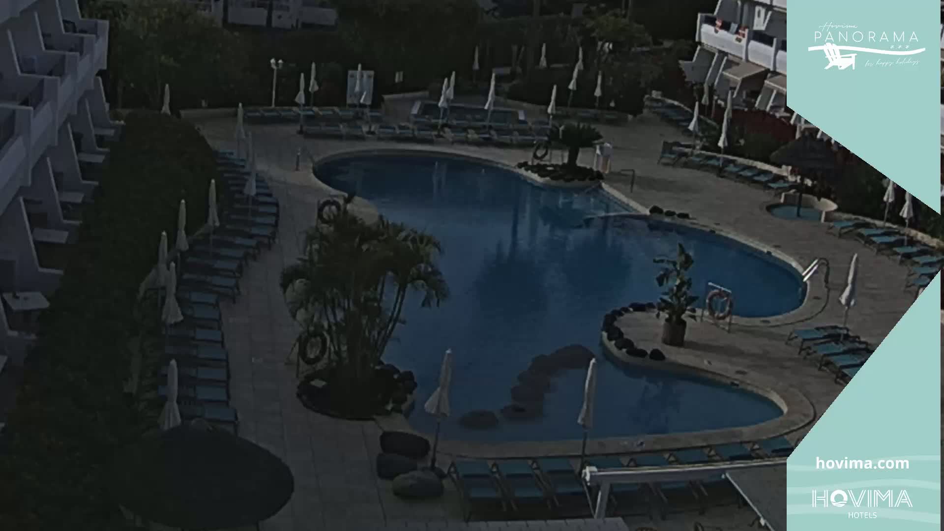 An outdoor swimming pool with surrounding lounge chairs and umbrellas is nestled between buildings under a dimly lit sky.