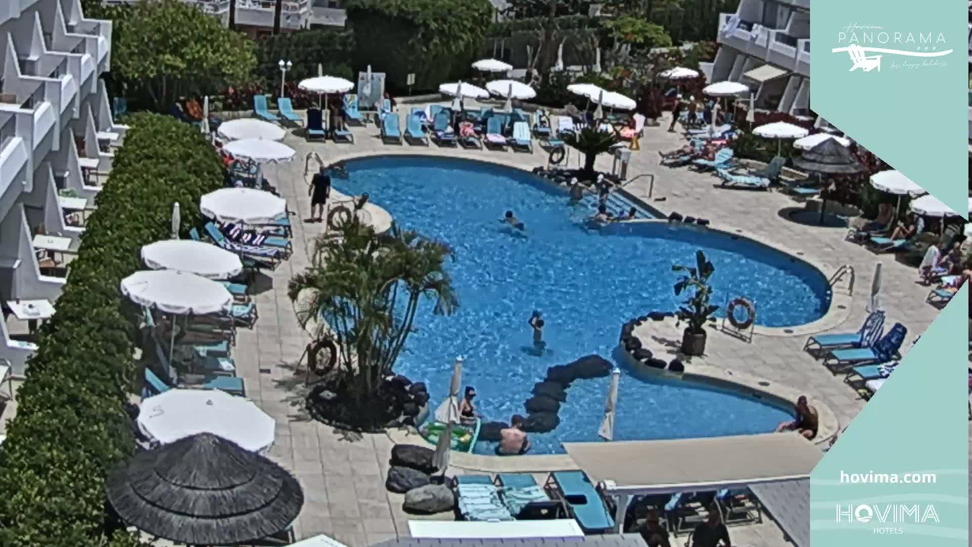 A resort pool area with several people swimming and lounging on sunny day, surrounded by white umbrellas, lounge chairs, and low-rise buildings.