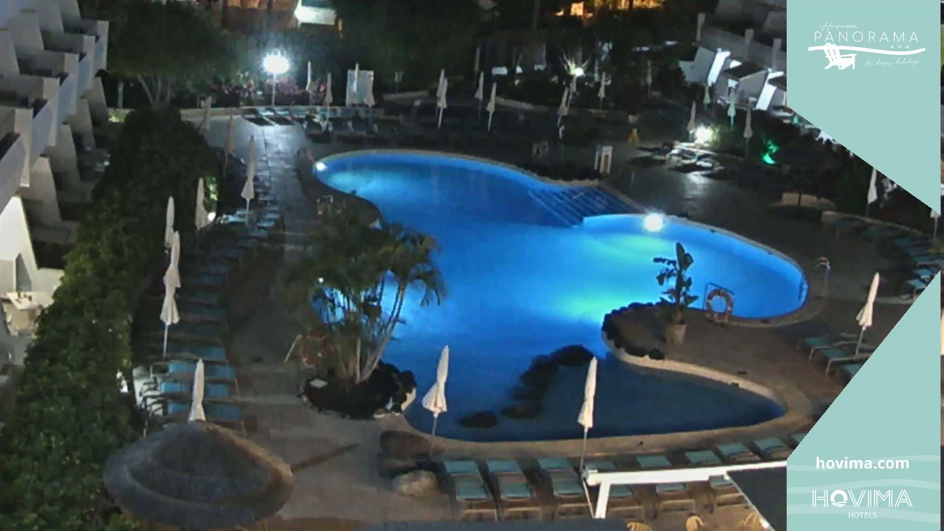 A nighttime aerial view of a kidney-shaped swimming pool surrounded by lounge chairs and palm trees at a resort.