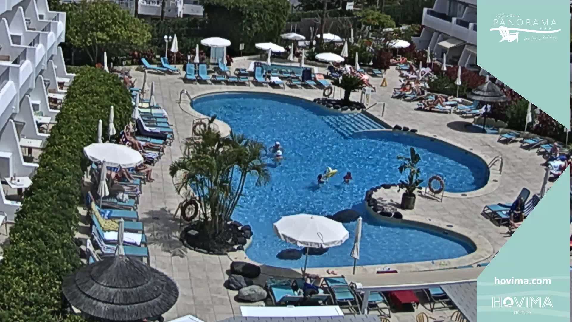An outdoor swimming pool surrounded by lounge chairs and umbrellas, with people relaxing and swimming on a sunny day.