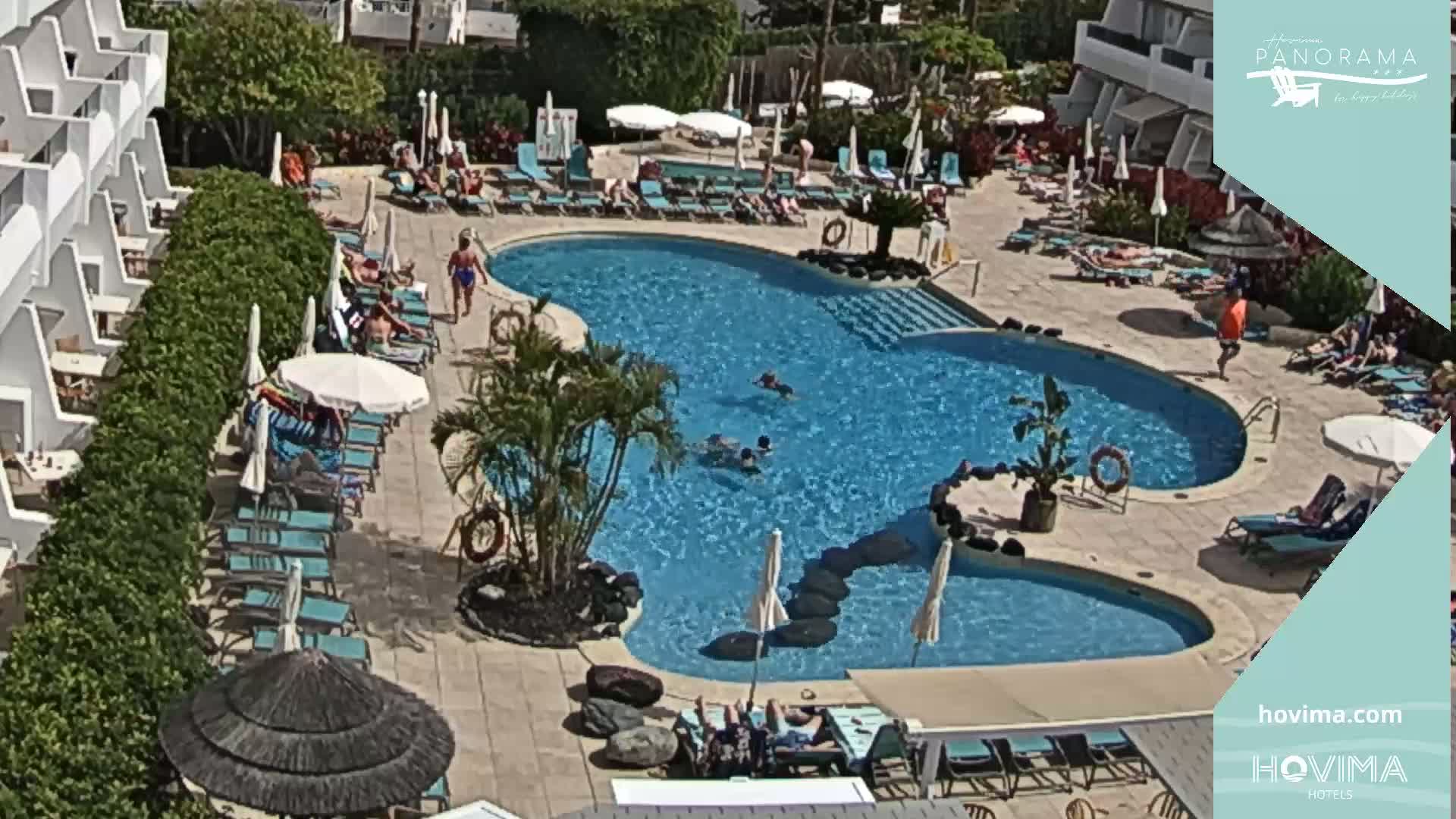 An outdoor swimming pool surrounded by lounge chairs and umbrellas, with people swimming and relaxing in the sunny weather.