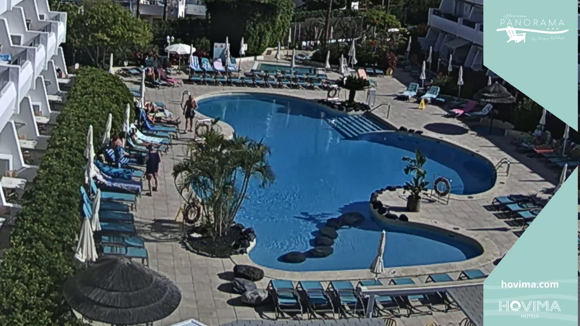 A sunny outdoor scene shows a kidney-shaped swimming pool surrounded by lounge chairs and umbrellas, with people relaxing around the pool area near a white building.