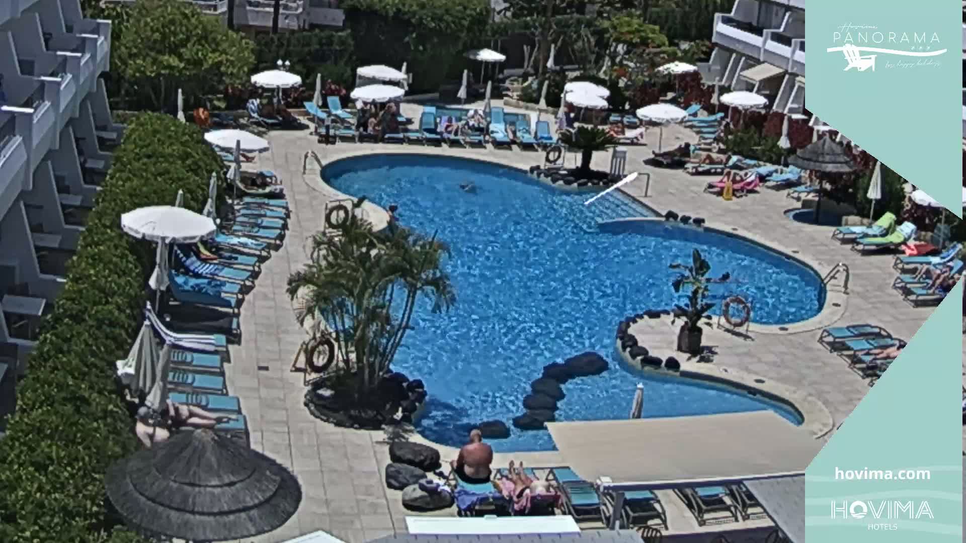 An outdoor swimming pool surrounded by lounge chairs and umbrellas, with people sunbathing and swimming on a sunny day.