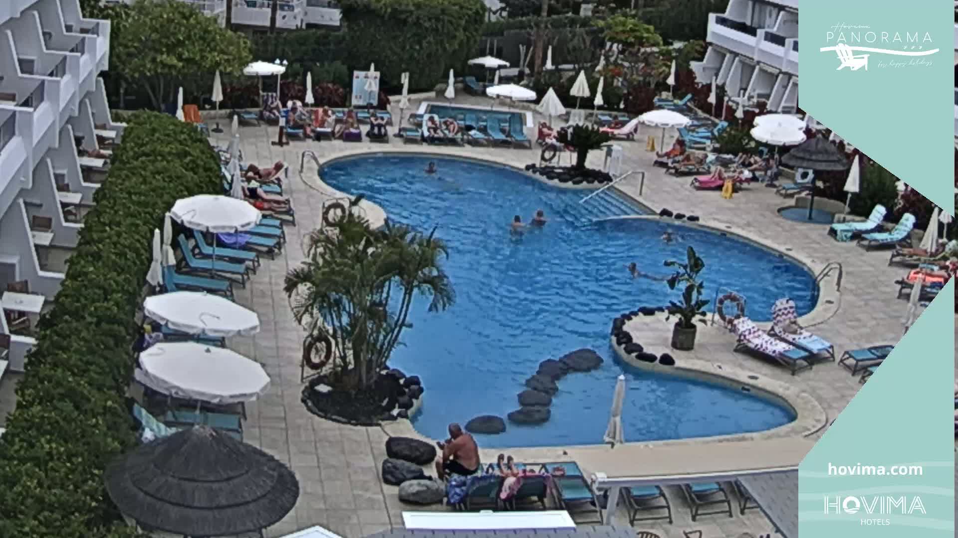 An outdoor swimming pool with many lounge chairs and umbrellas surrounding it, and a building in the background, on a sunny day.