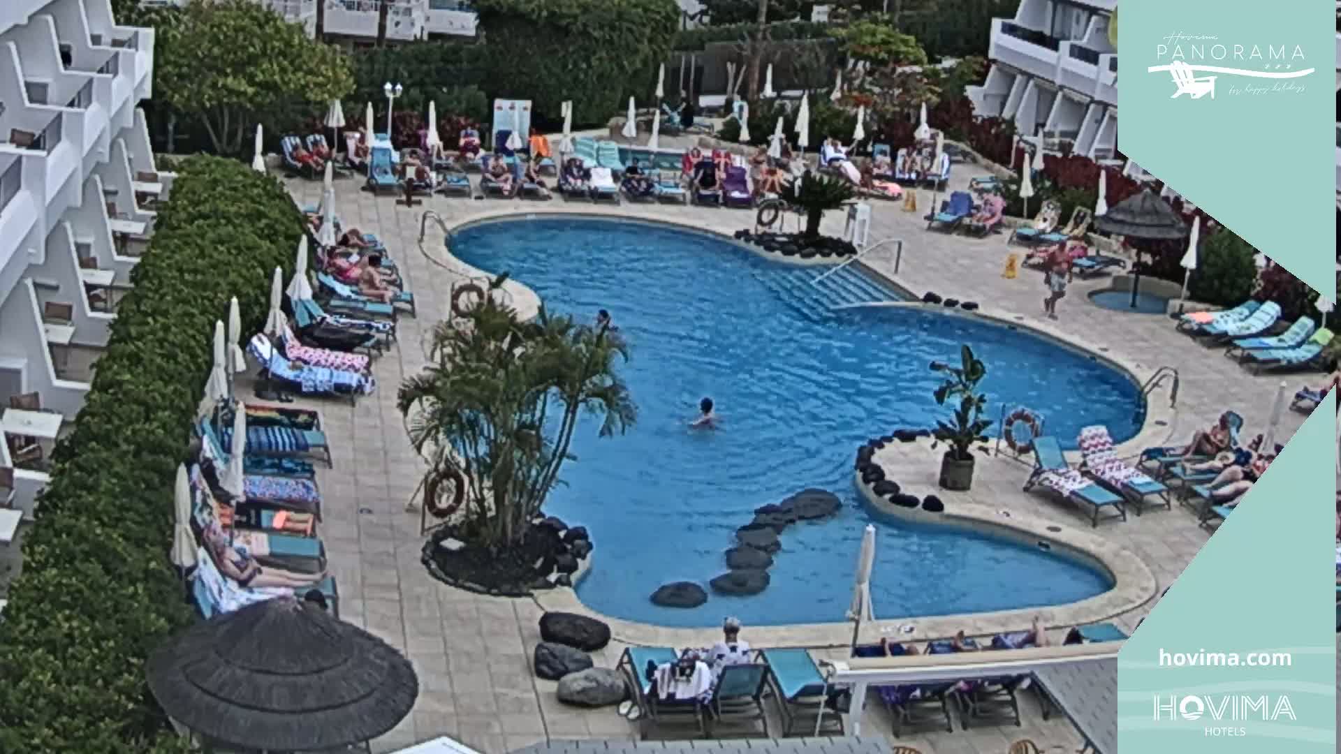 An outdoor pool with many people lounging on surrounding chairs and a few swimming, in sunny weather.