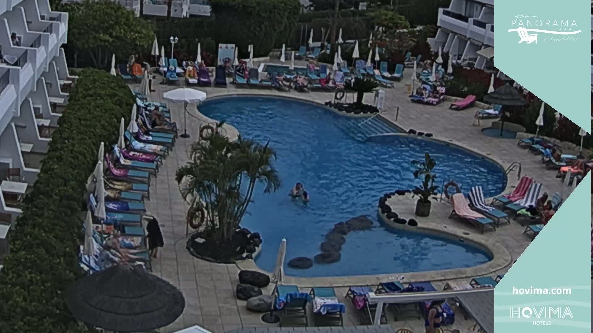 An outdoor pool surrounded by lounge chairs and umbrellas, with people relaxing and swimming, on a partly sunny day.