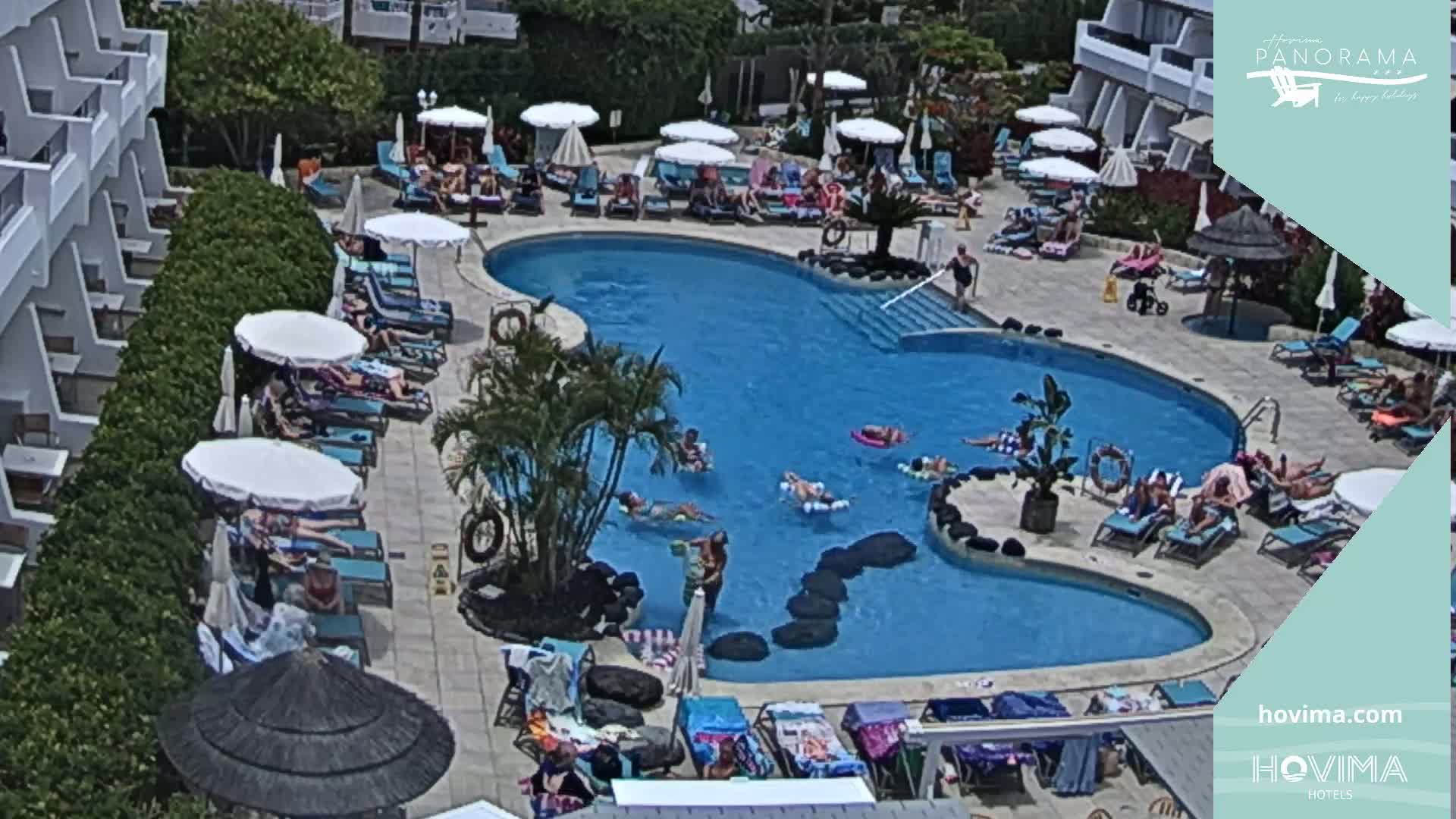 A sunny outdoor scene shows a kidney-shaped swimming pool surrounded by sunbathers lounging on chairs under umbrellas near a hotel.