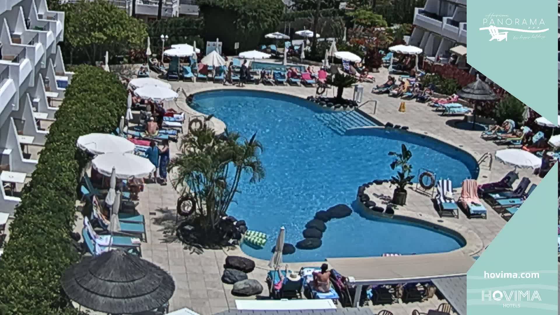 An aerial view of a resort pool area on a sunny day shows a large, irregularly shaped swimming pool surrounded by lounge chairs, umbrellas, and people relaxing.