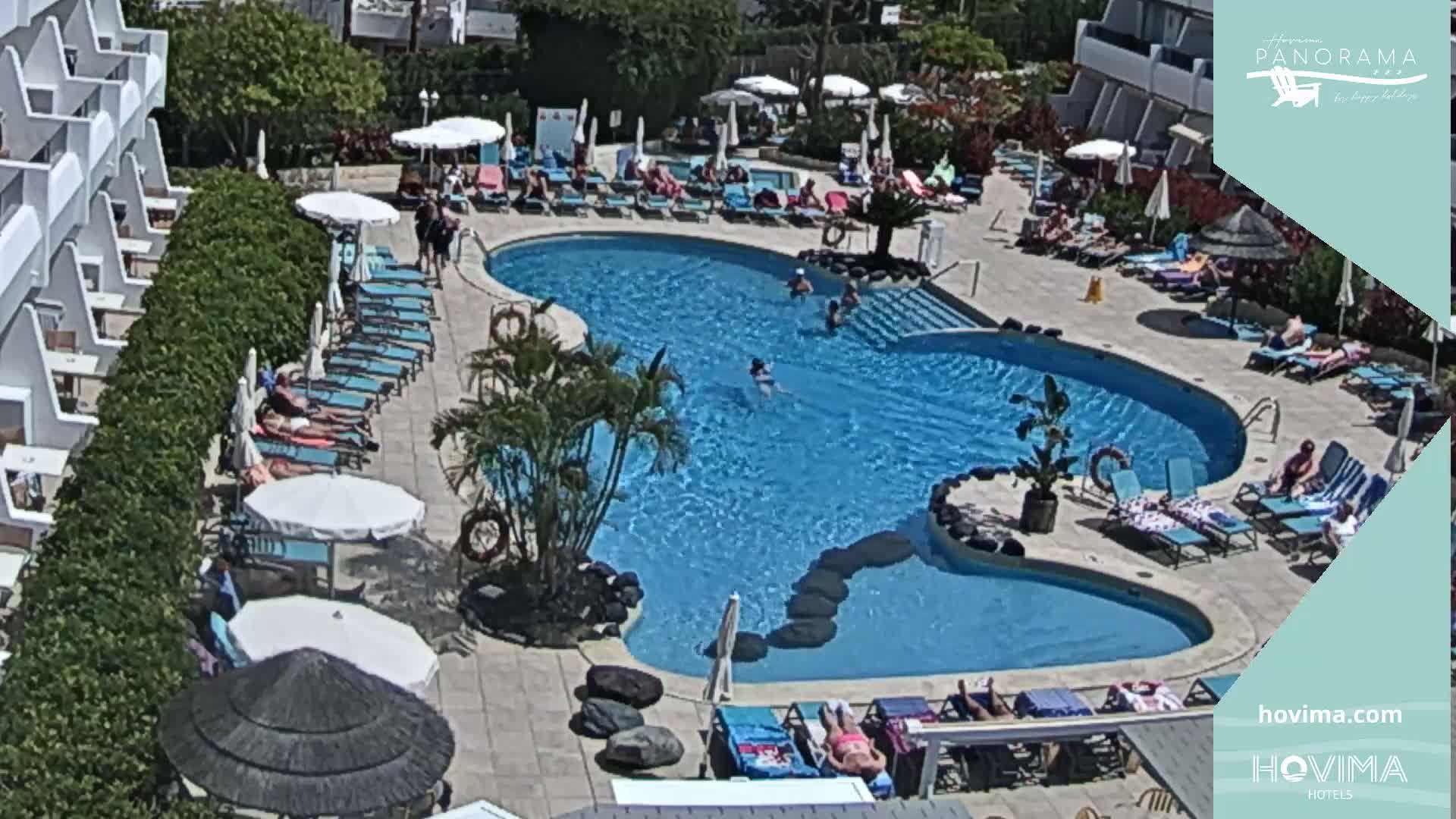 An aerial view of a resort pool area shows a large, kidney-shaped swimming pool surrounded by lounge chairs and umbrellas under a sunny sky.