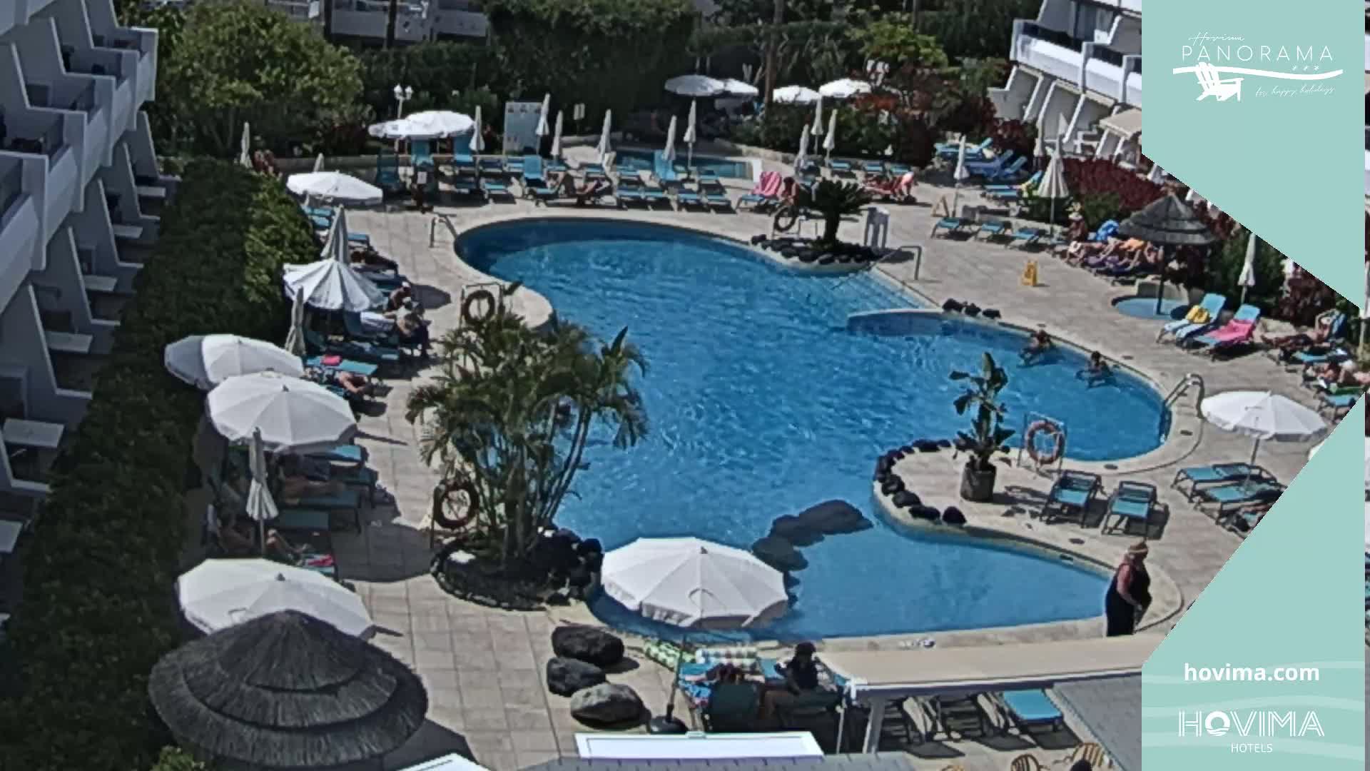 A large, kidney-shaped swimming pool surrounded by lounge chairs, umbrellas, and people relaxing on a sunny day.