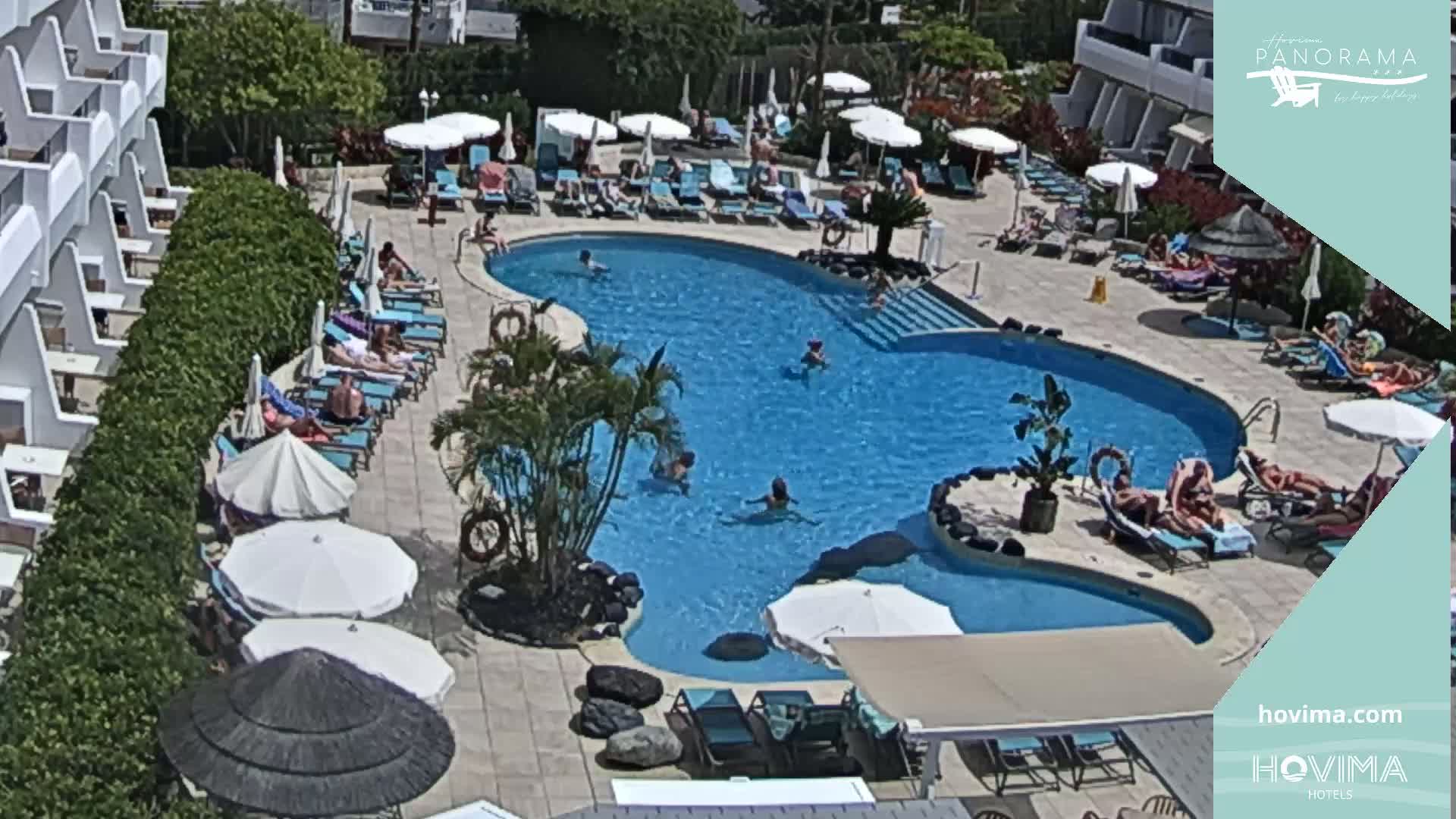 An outdoor pool surrounded by lounge chairs and umbrellas, with people swimming and sunbathing in sunny weather.