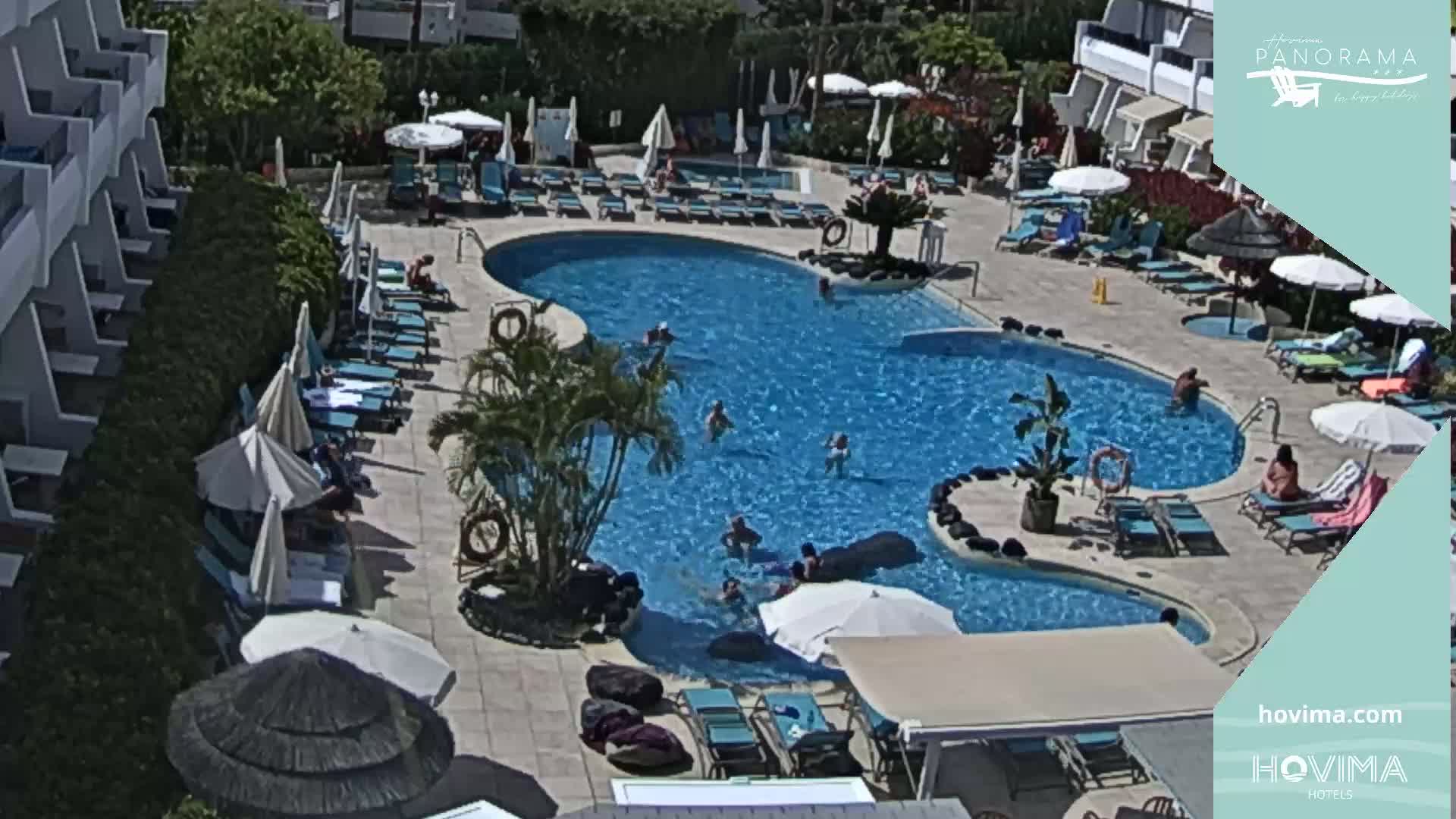 An outdoor pool surrounded by lounge chairs, umbrellas, and buildings on a sunny day.