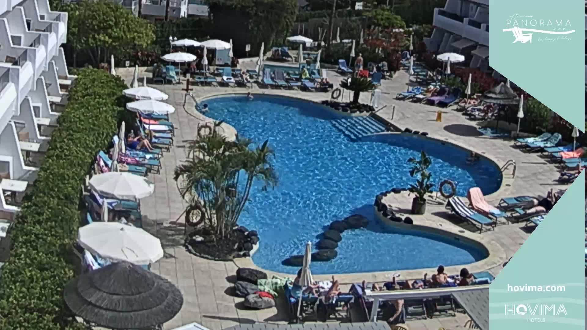 A sunny outdoor scene shows a kidney-shaped swimming pool surrounded by lounge chairs, umbrellas, and people relaxing around the pool area near a hotel building.