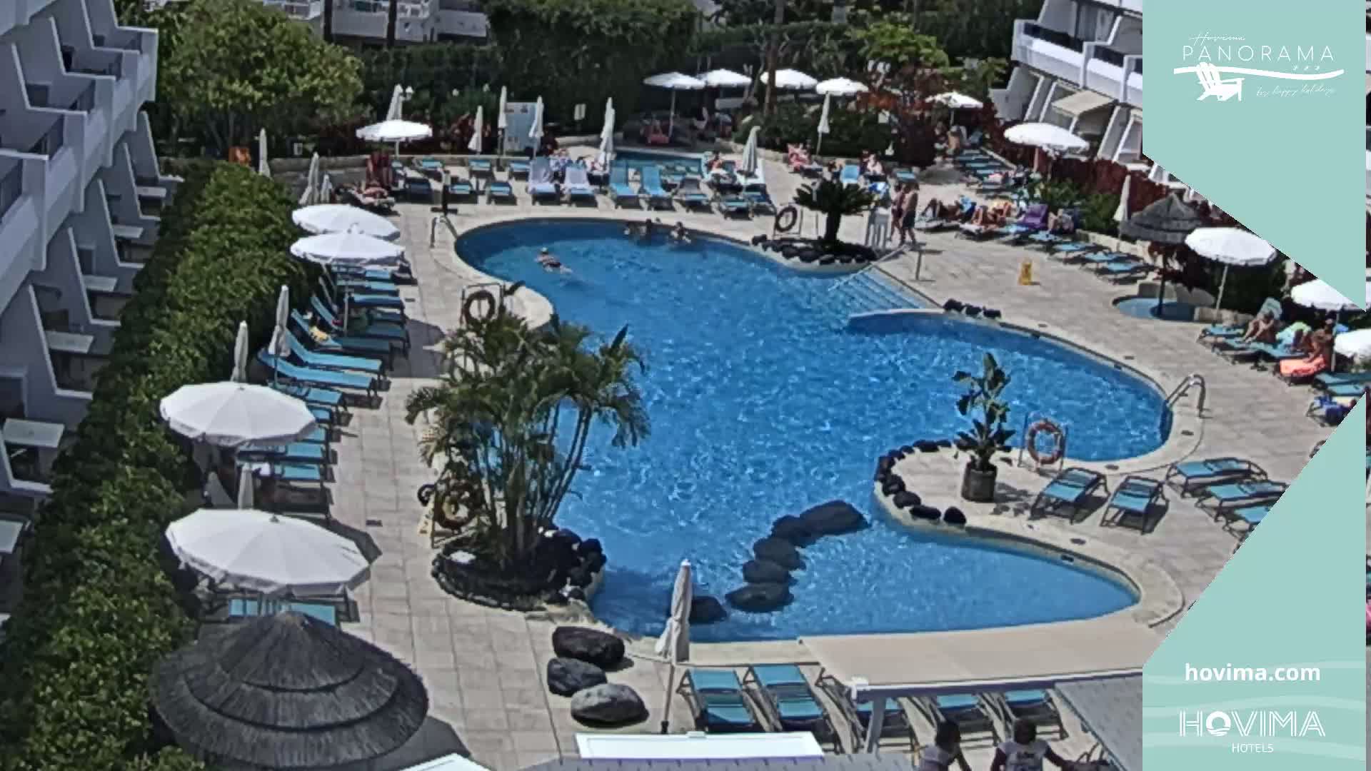 An outdoor pool surrounded by lounge chairs and umbrellas, with several people swimming and sunbathing on a sunny day.