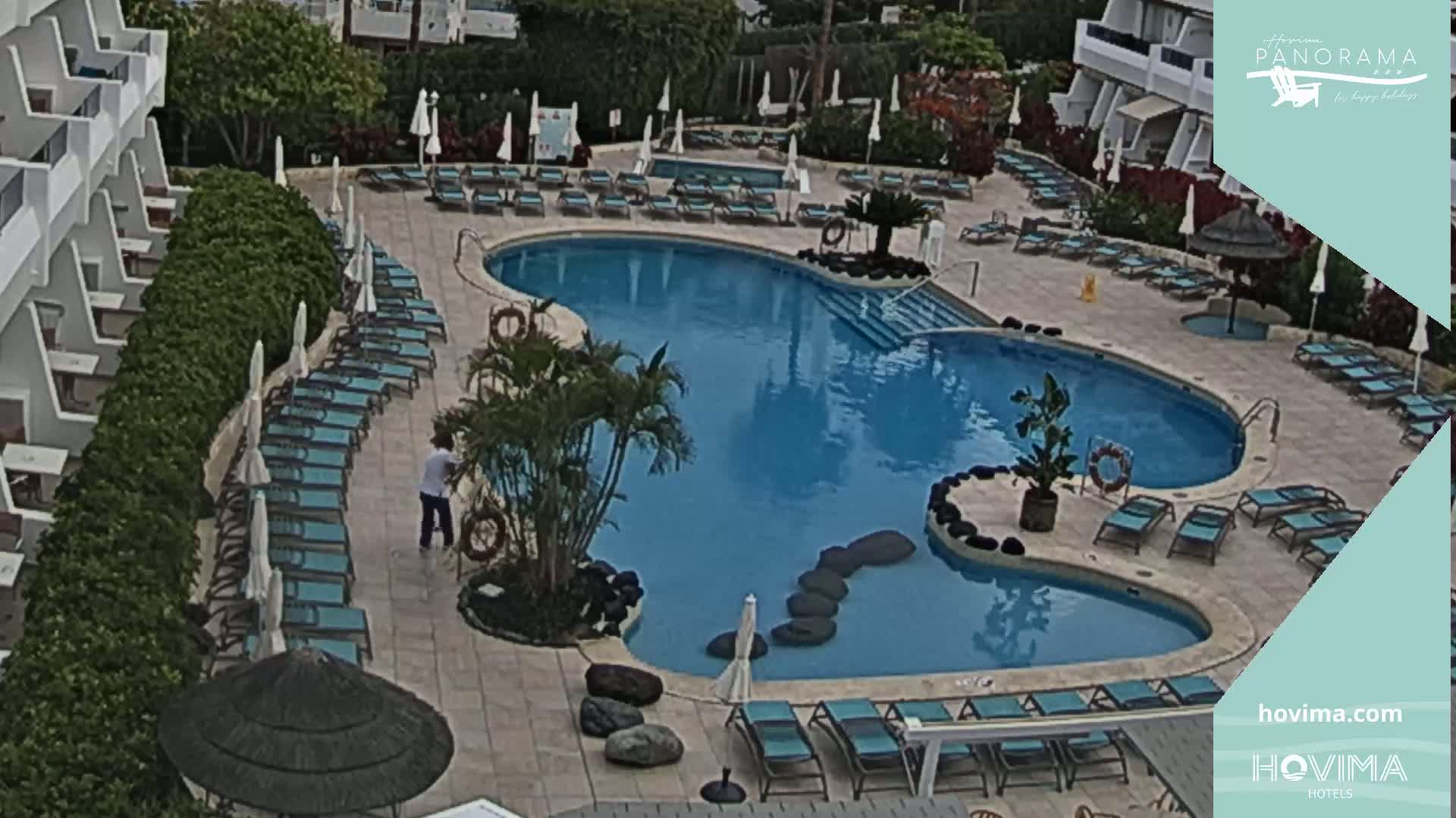 An outdoor pool surrounded by lounge chairs and umbrellas, situated next to a multi-story building, under a mostly sunny sky.
