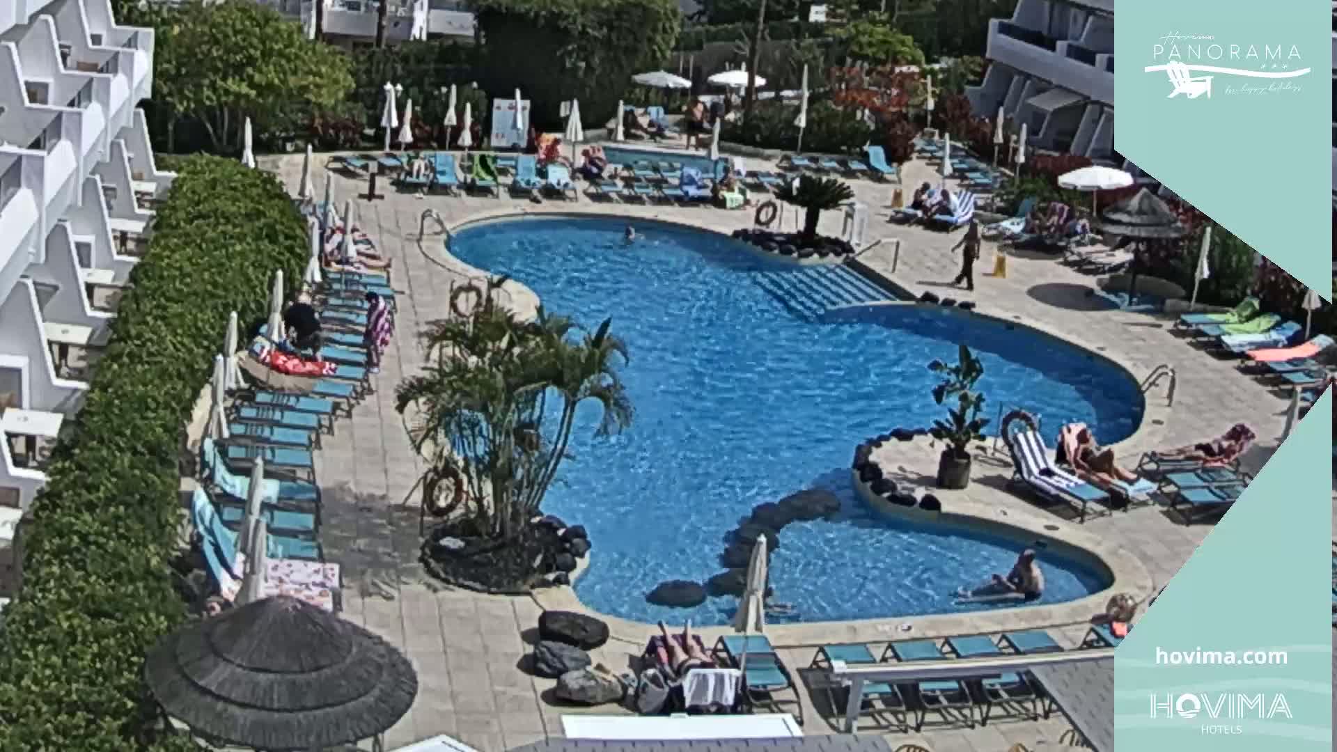 An aerial view shows a sunny outdoor pool area with many lounge chairs surrounding a kidney-shaped pool, some people are swimming and sunbathing.