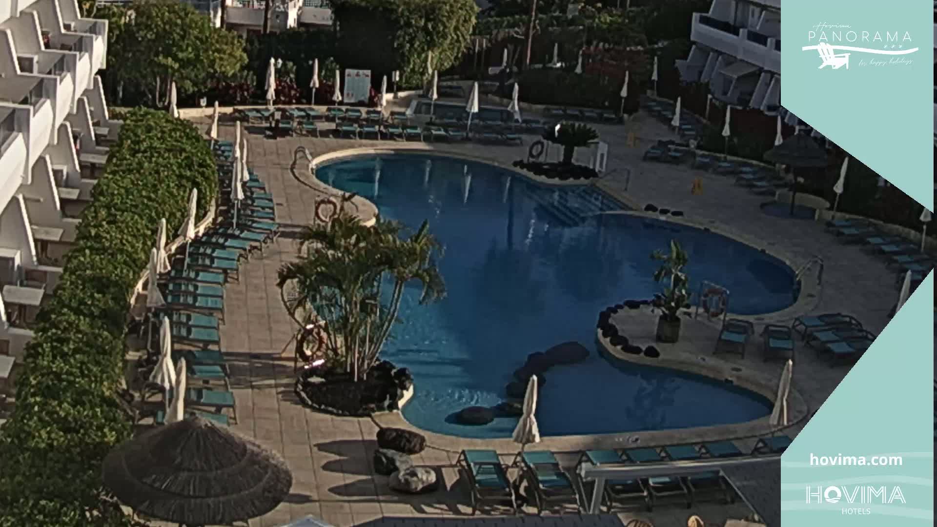 An outdoor pool with surrounding lounge chairs and umbrellas is nestled between a white building and lush greenery under a sunny sky.