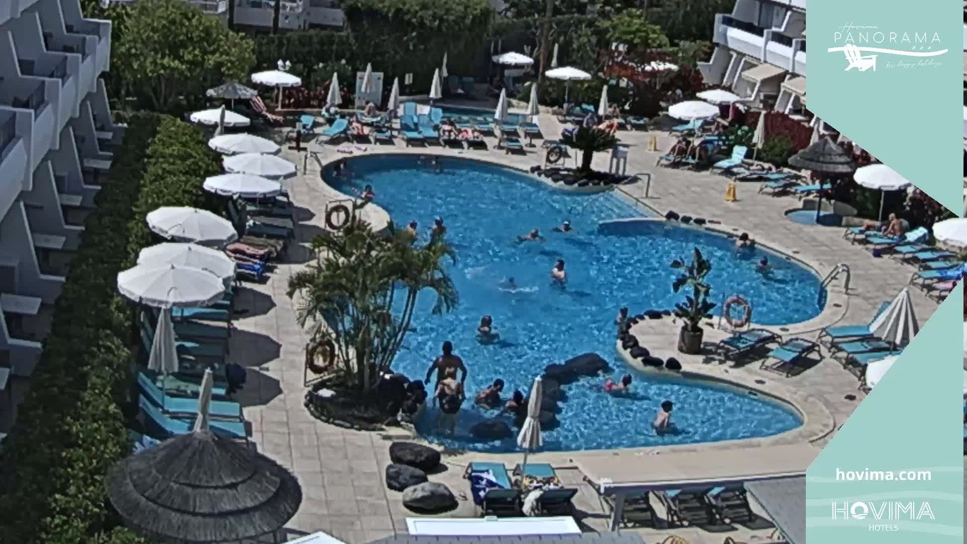 A large, irregularly shaped swimming pool surrounded by lounge chairs, umbrellas, and palm trees, with many people swimming and sunbathing on a sunny day.