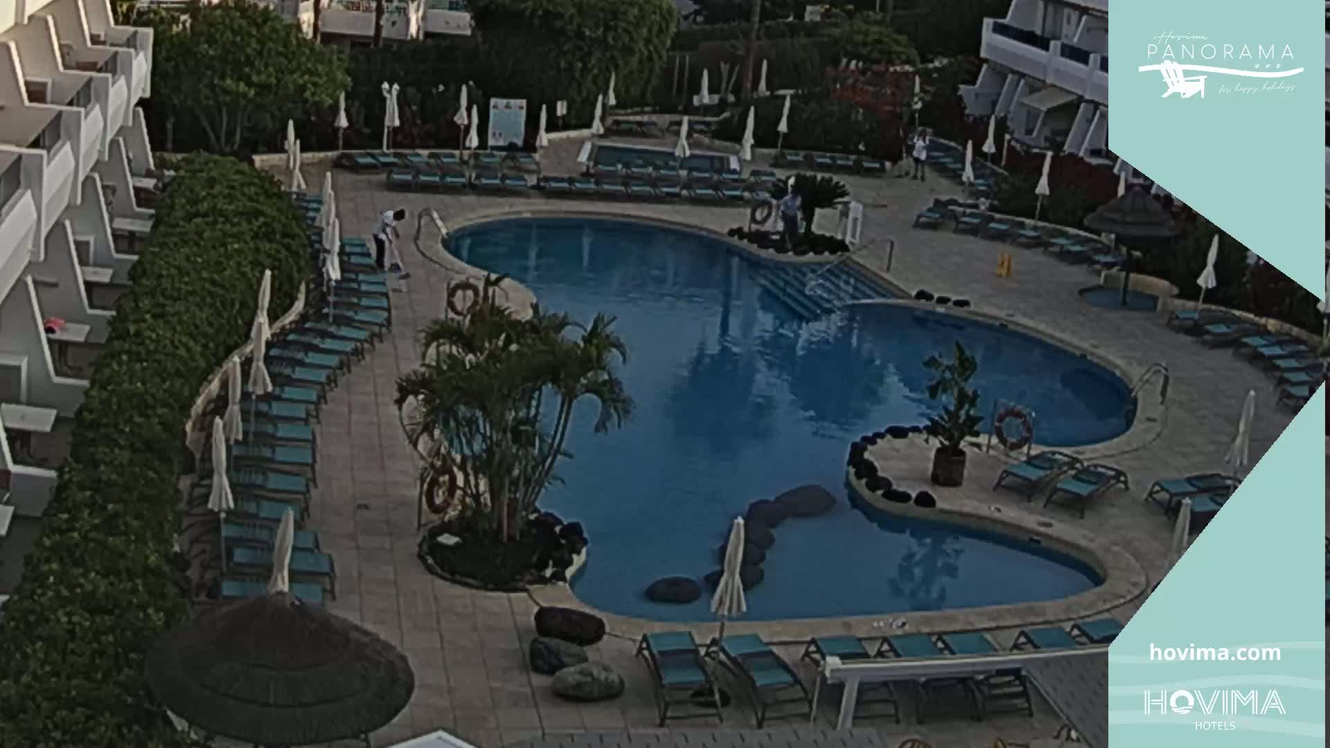 An outdoor swimming pool surrounded by lounge chairs and umbrellas at a resort, with a person sweeping near the pool.