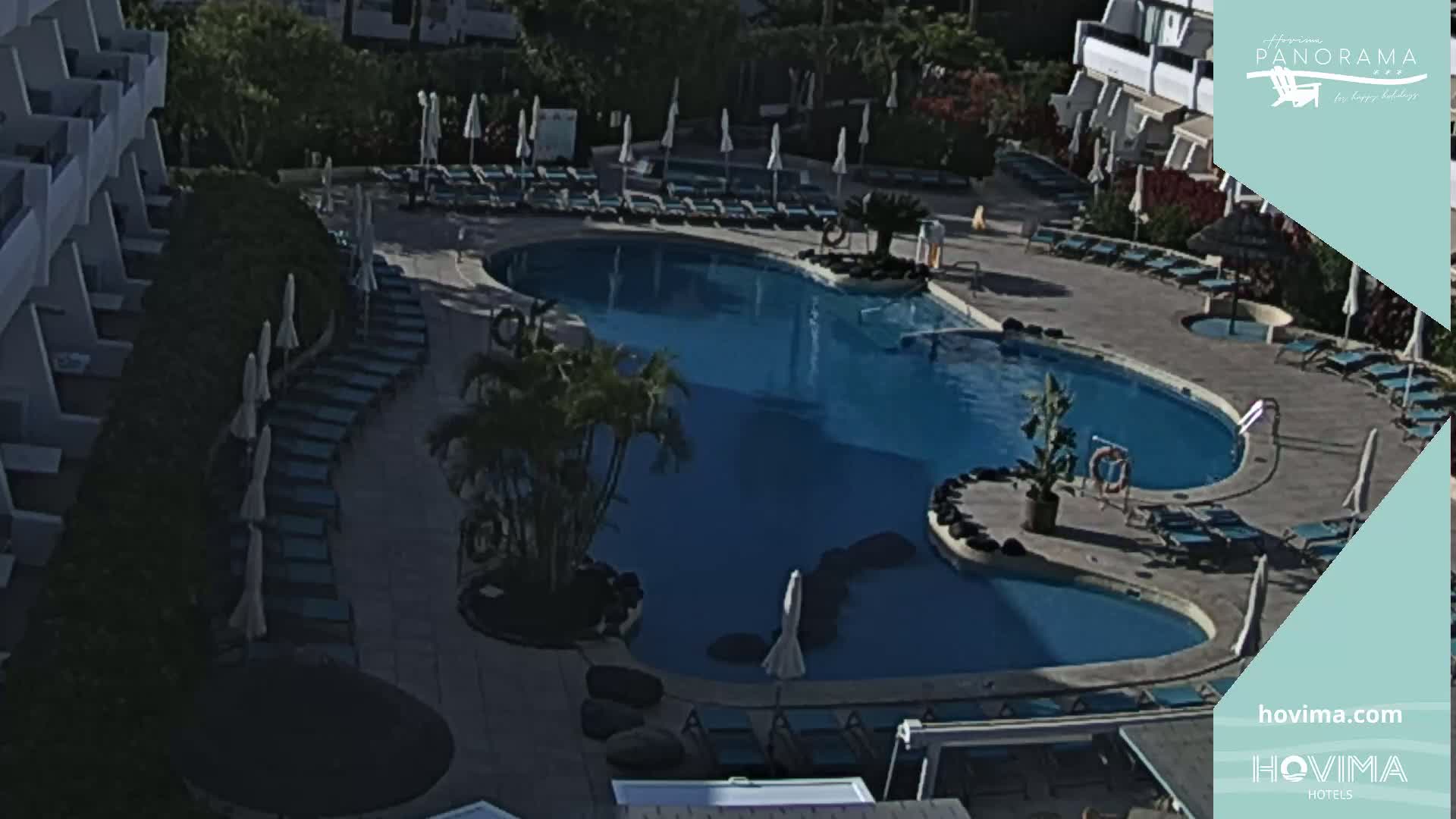 An outdoor pool with surrounding lounge chairs and umbrellas, situated next to a building, on a sunny day.