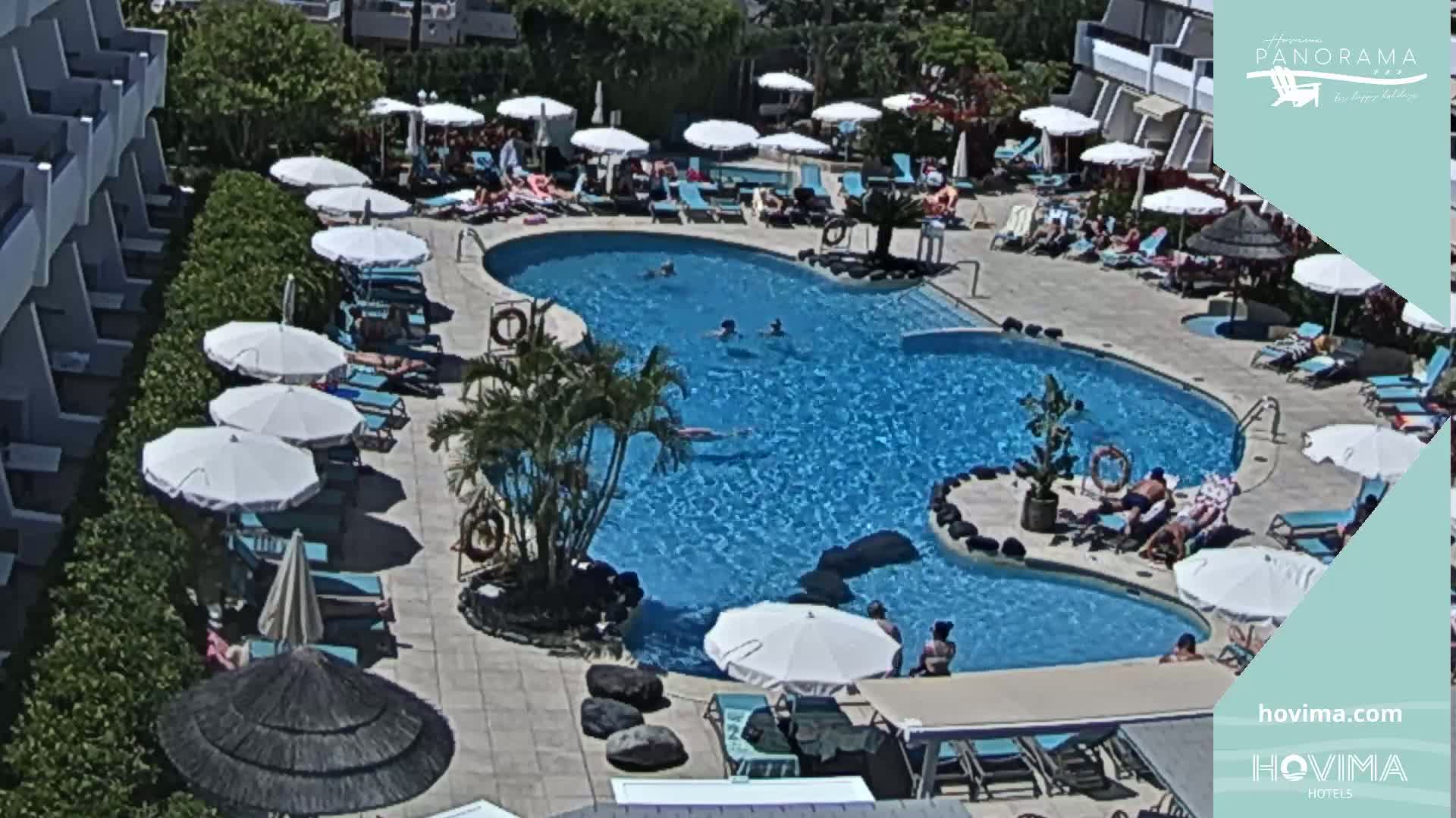 A sunny outdoor scene shows a swimming pool surrounded by lounge chairs, umbrellas, and a building, with several people swimming and sunbathing.
