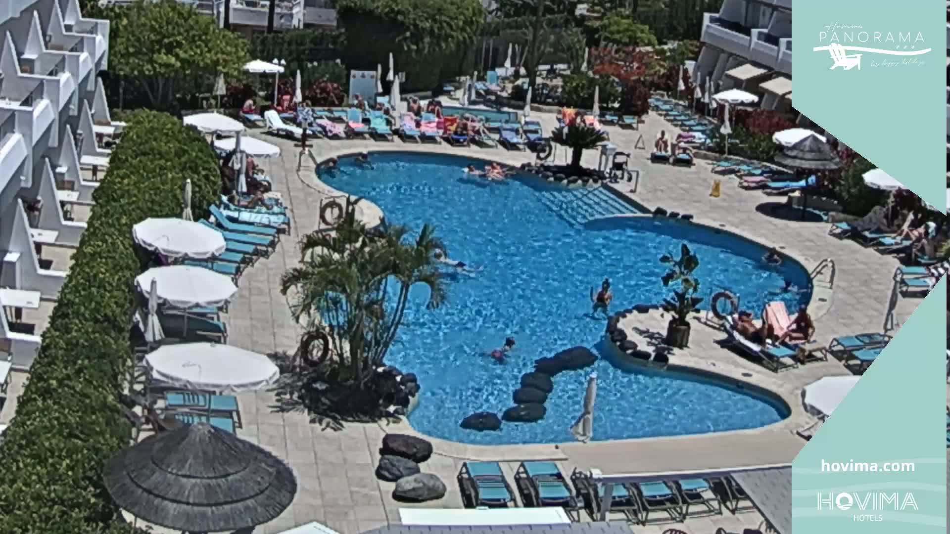 An outdoor swimming pool surrounded by lounge chairs and umbrellas, with people swimming and sunbathing on a sunny day.