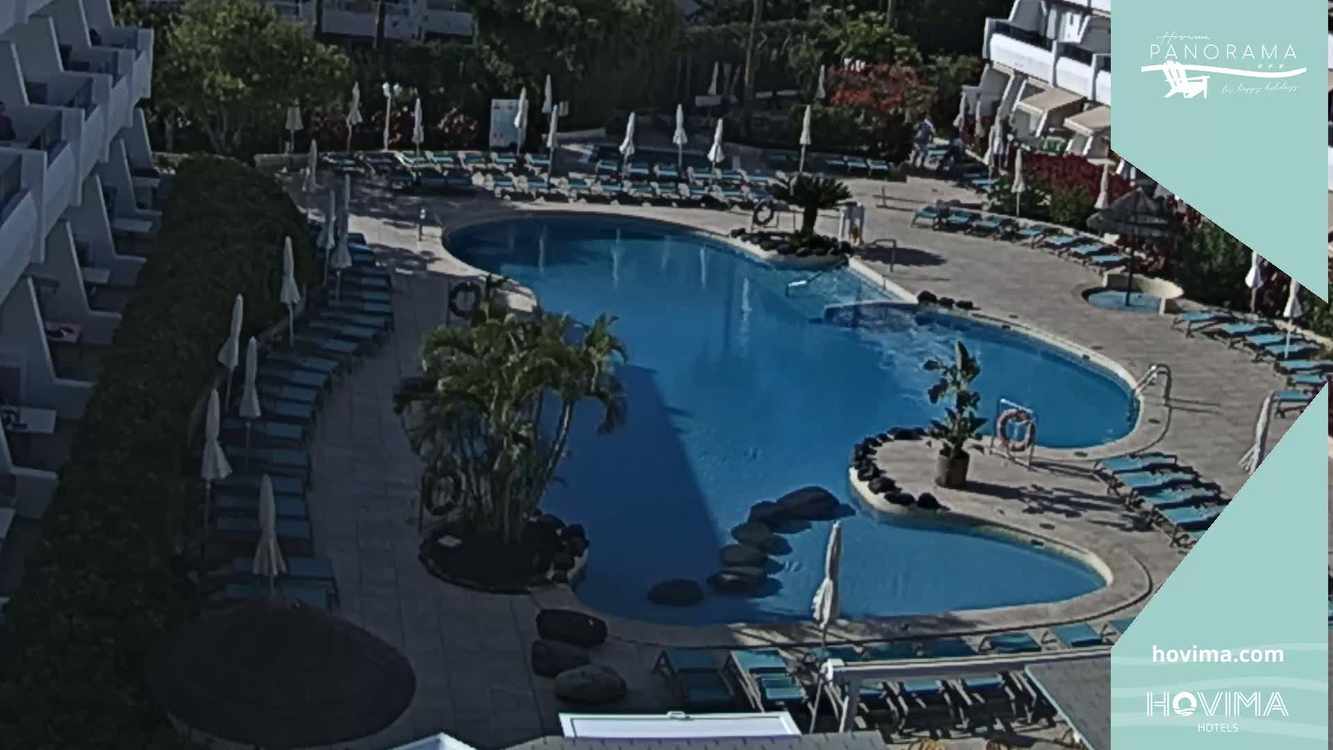 An outdoor pool with surrounding lounge chairs and umbrellas, situated beside a building under sunny conditions.