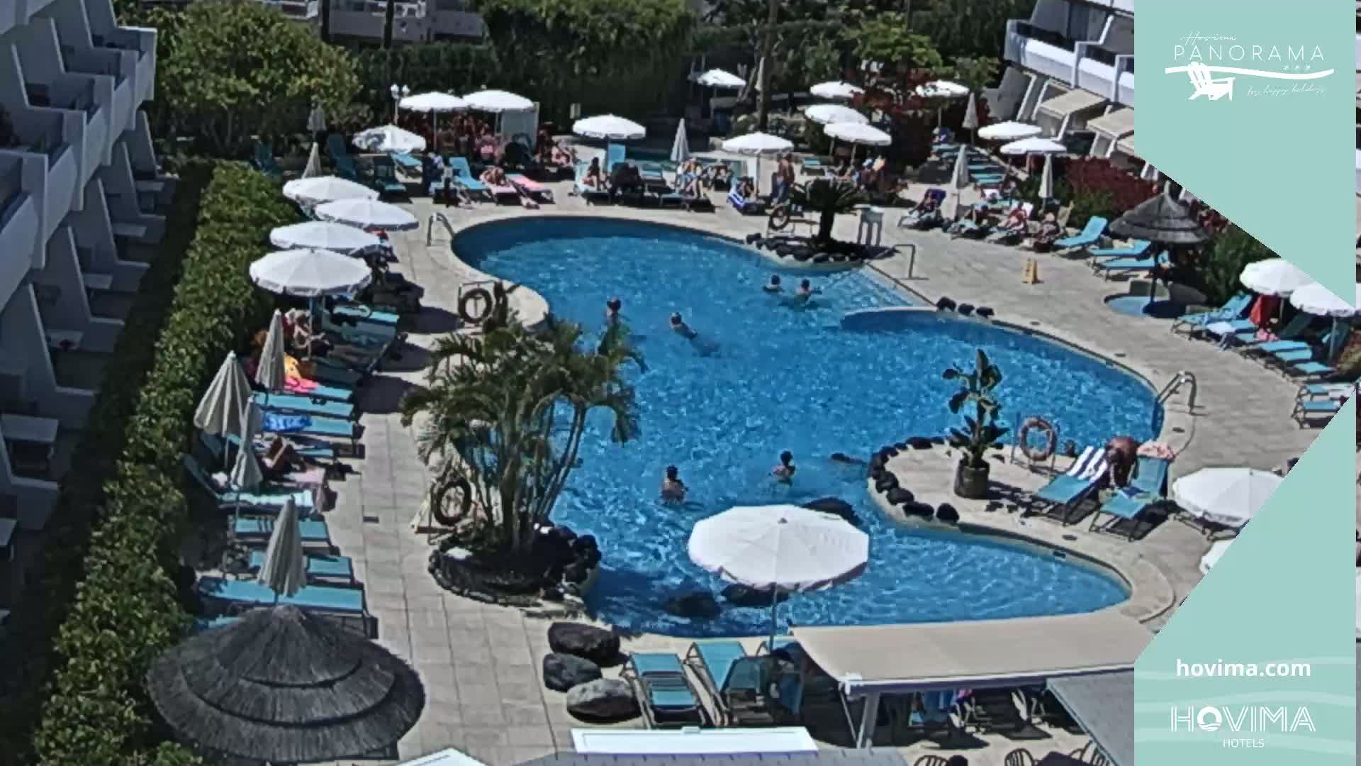 An outdoor pool surrounded by lounge chairs and umbrellas, with people swimming and sunbathing on a sunny day.