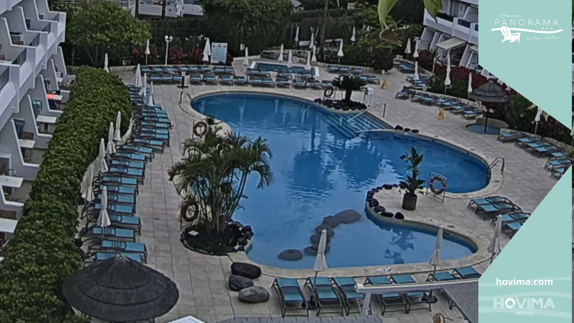An outdoor pool with many lounge chairs and umbrellas surrounding it, situated beside a multi-story building.