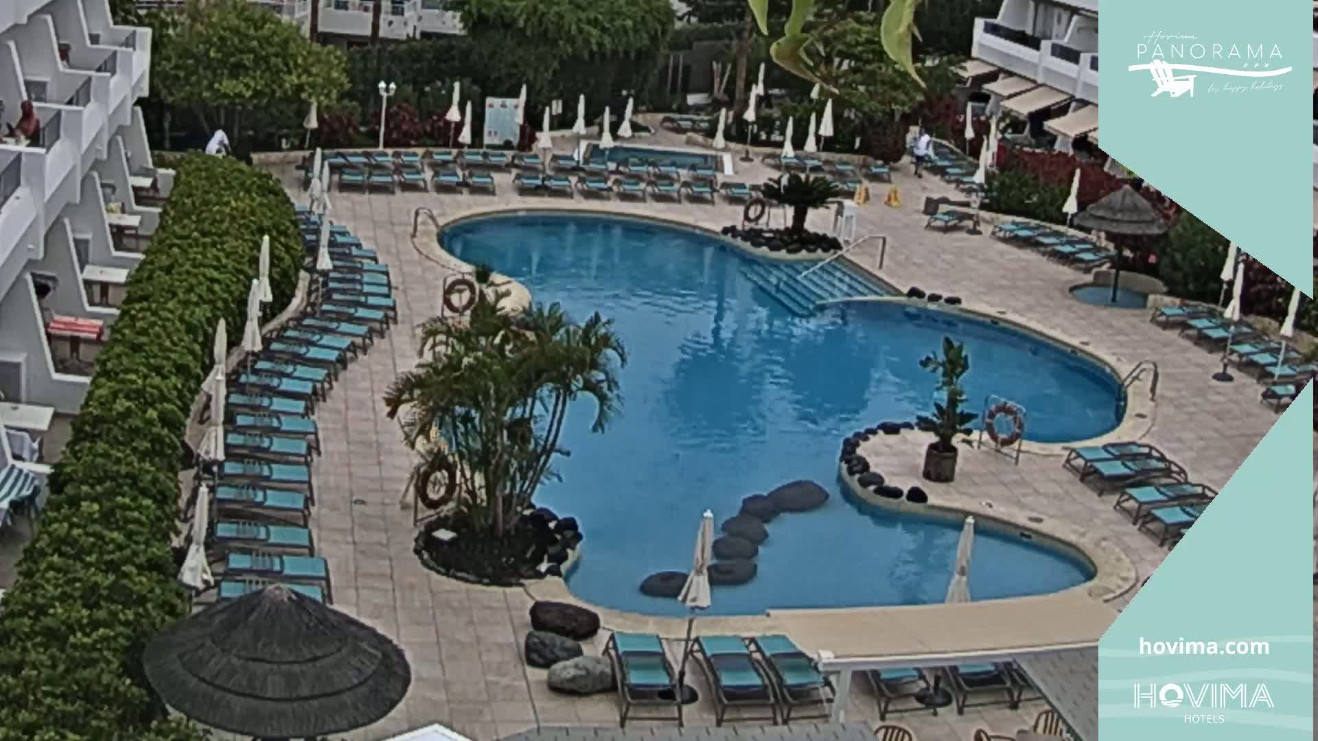An outdoor pool with surrounding lounge chairs and umbrellas is situated next to a multi-story building.