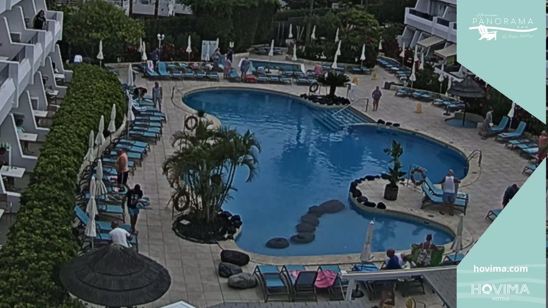 An outdoor pool area with many lounge chairs and people, surrounded by a building and landscaping under a mostly sunny sky.