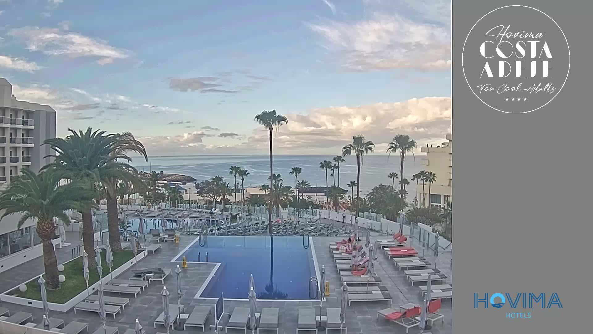 A resort pool area with lounge chairs and palm trees overlooks a calm ocean under a partly cloudy sky.
