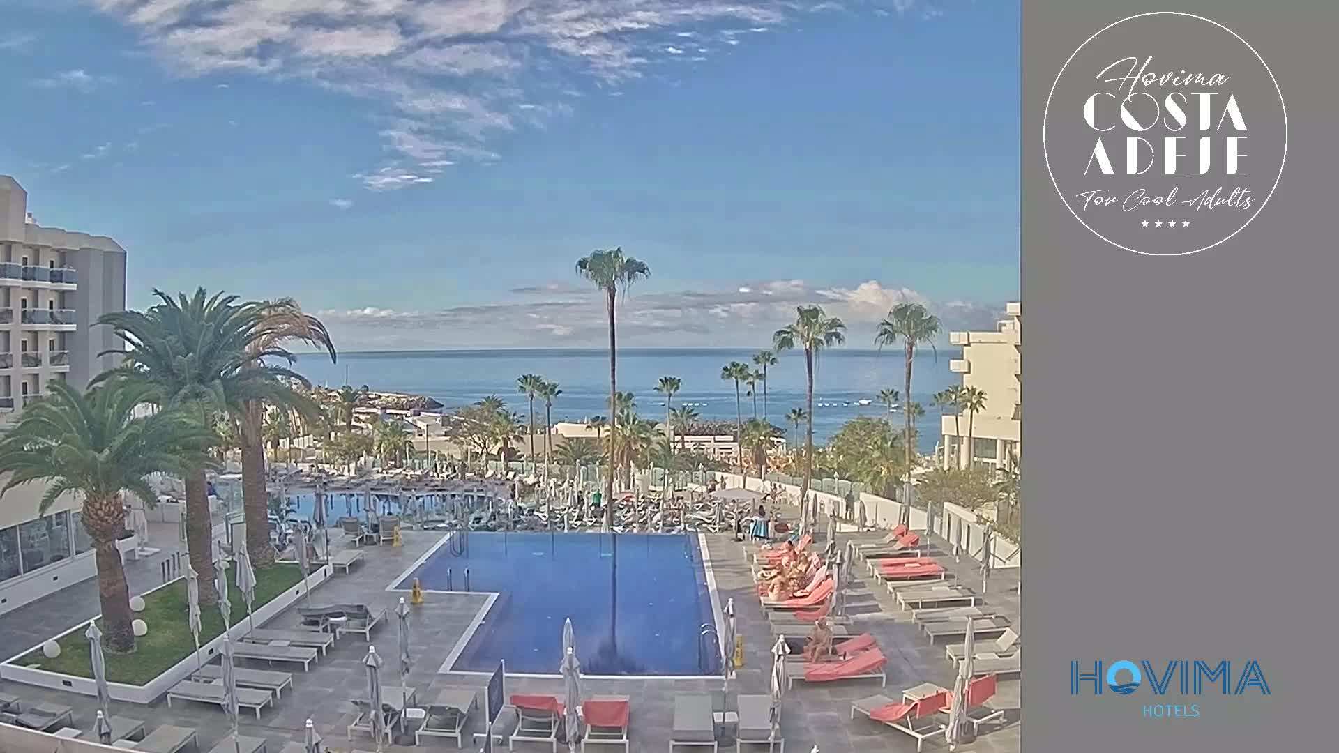 A resort pool area with lounge chairs and palm trees overlooks a calm ocean under a mostly sunny sky.