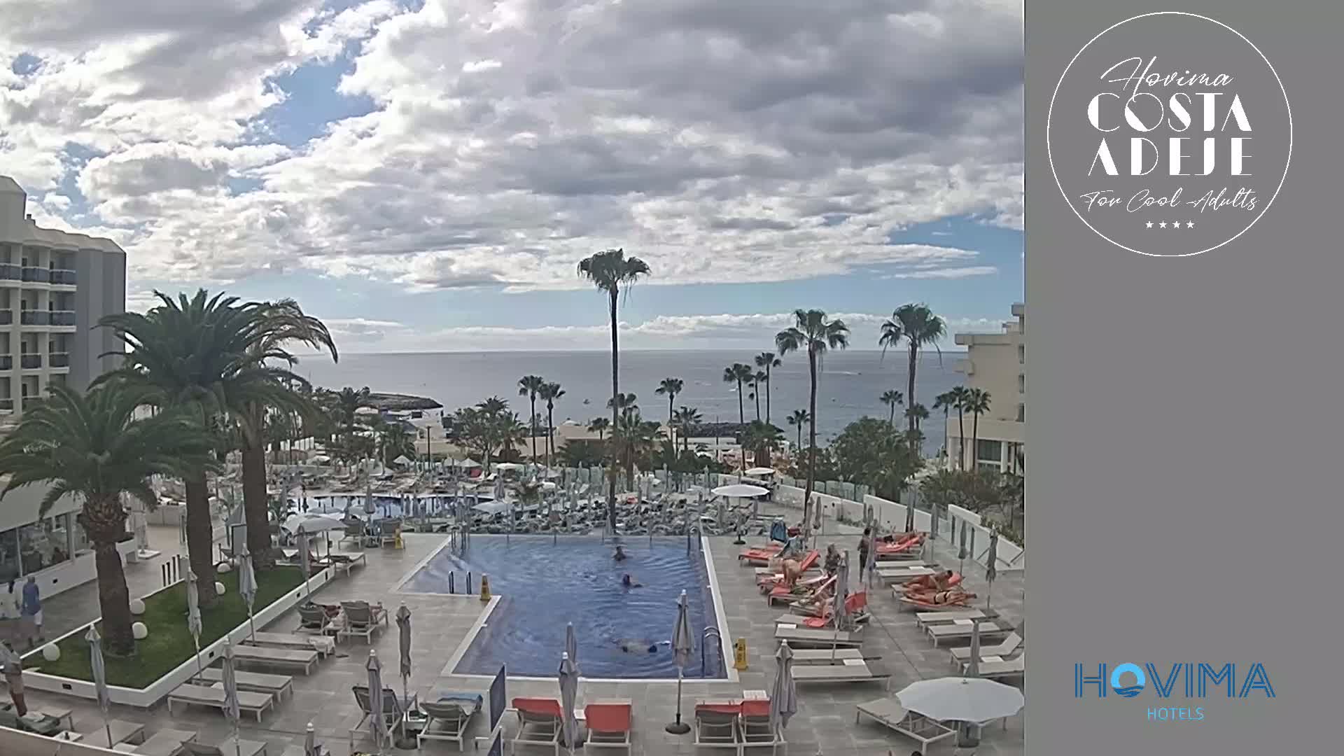 A resort pool with several people swimming and sunbathing is shown, overlooking a calm ocean under a partly cloudy sky.