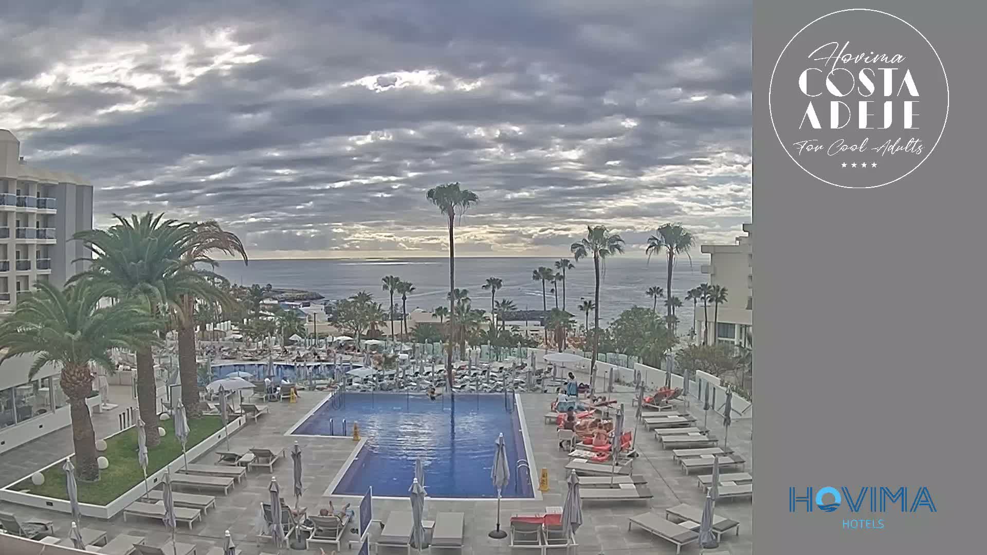 A resort pool area with lounge chairs and palm trees overlooks a calm ocean under a cloudy sky.
