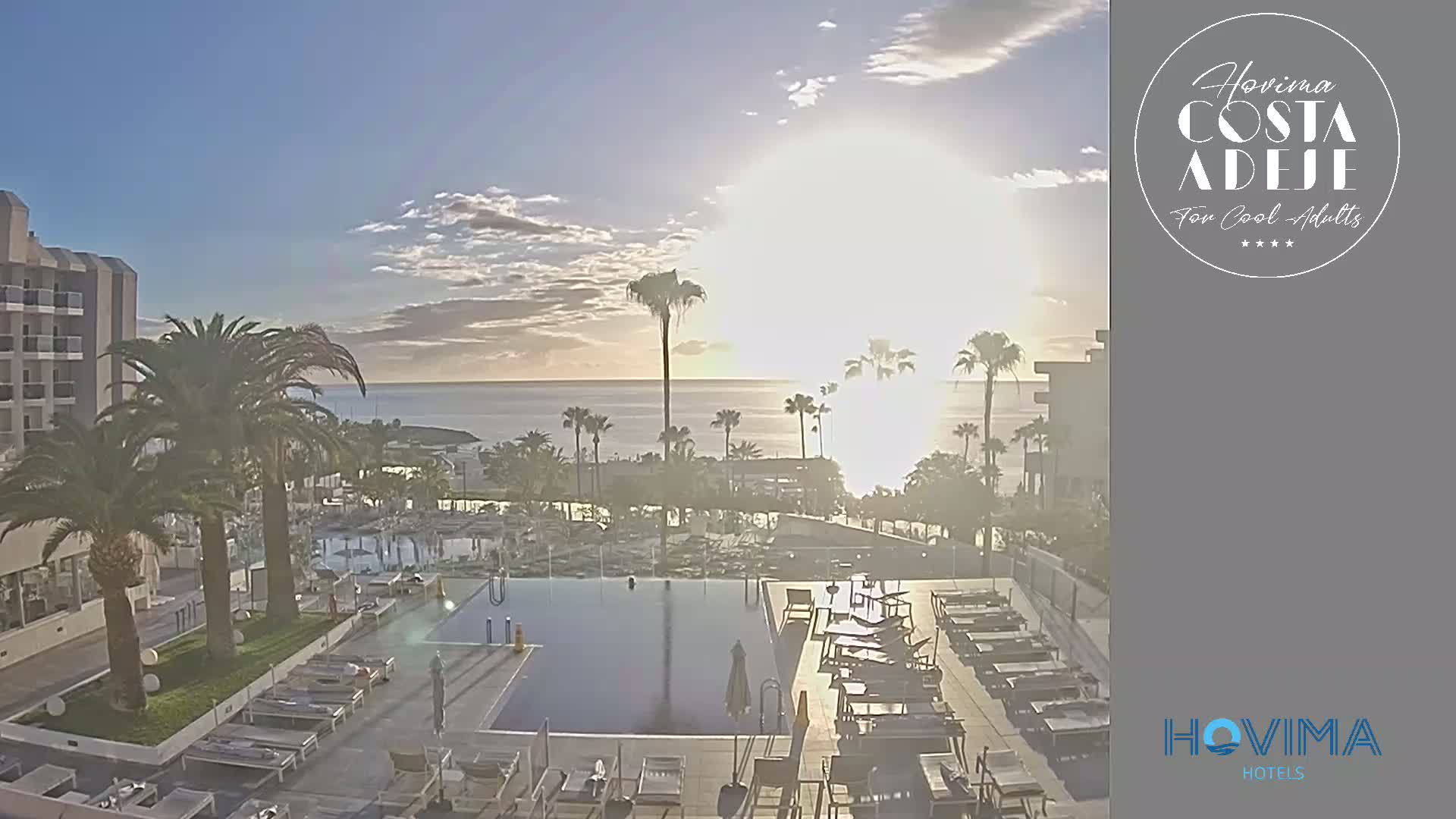 A resort pool area with palm trees and lounge chairs overlooks a calm ocean at sunrise.