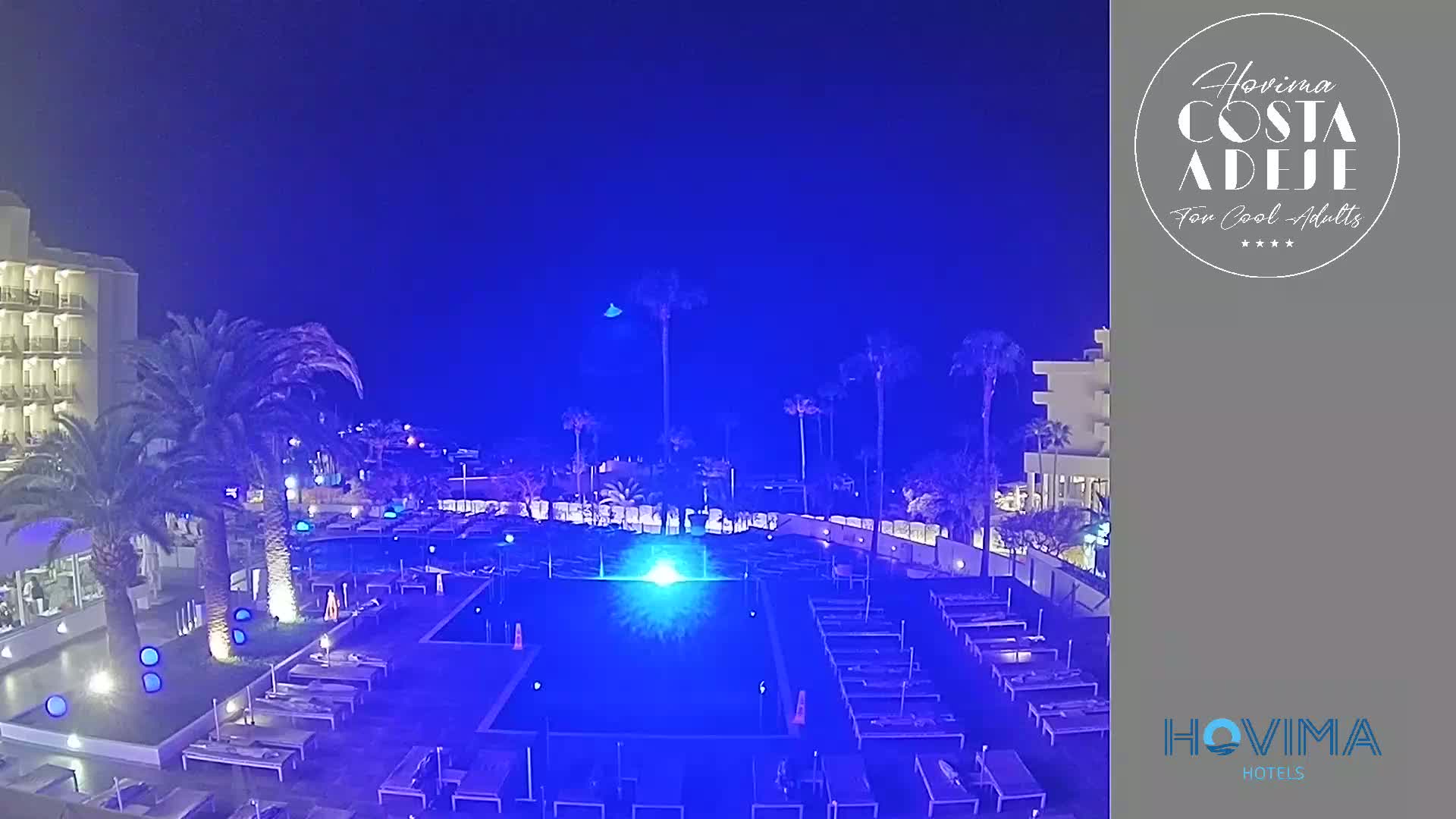 A nighttime view of a resort pool illuminated in blue, surrounded by palm trees, lounge chairs, and a multi-story hotel building under a clear, dark blue sky.