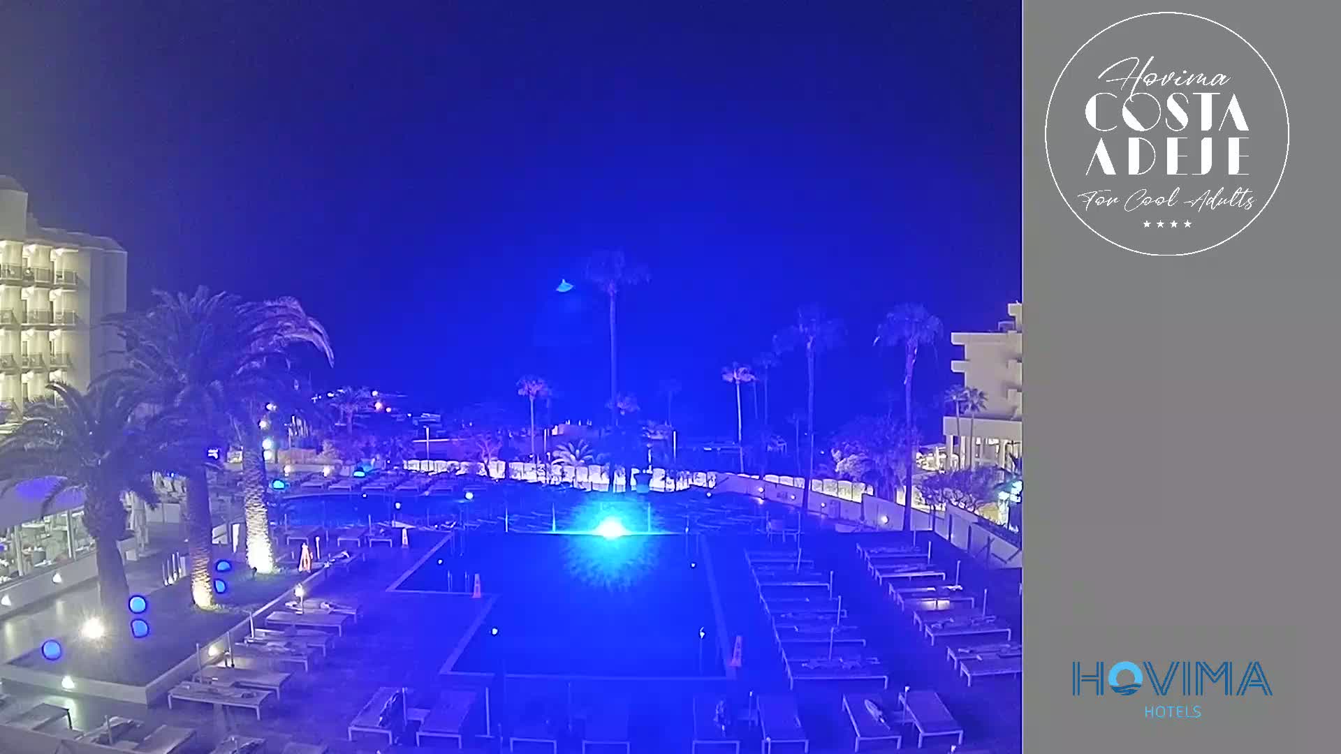 A nighttime view of a hotel pool area illuminated in blue, surrounded by palm trees and buildings under a clear, dark sky.