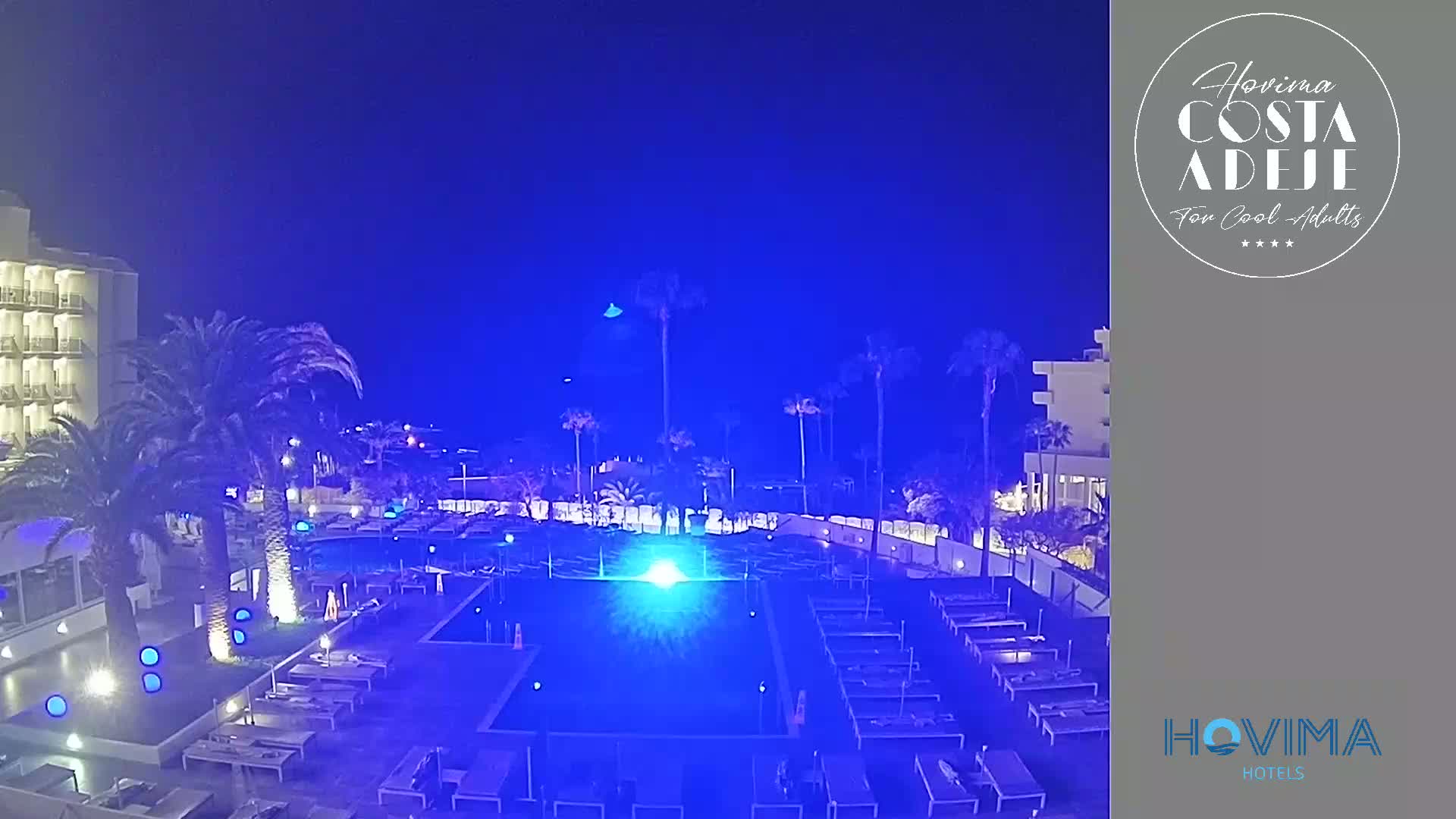 A resort pool area at night is illuminated in blue under a dark blue sky, surrounded by palm trees and a multi-story building.