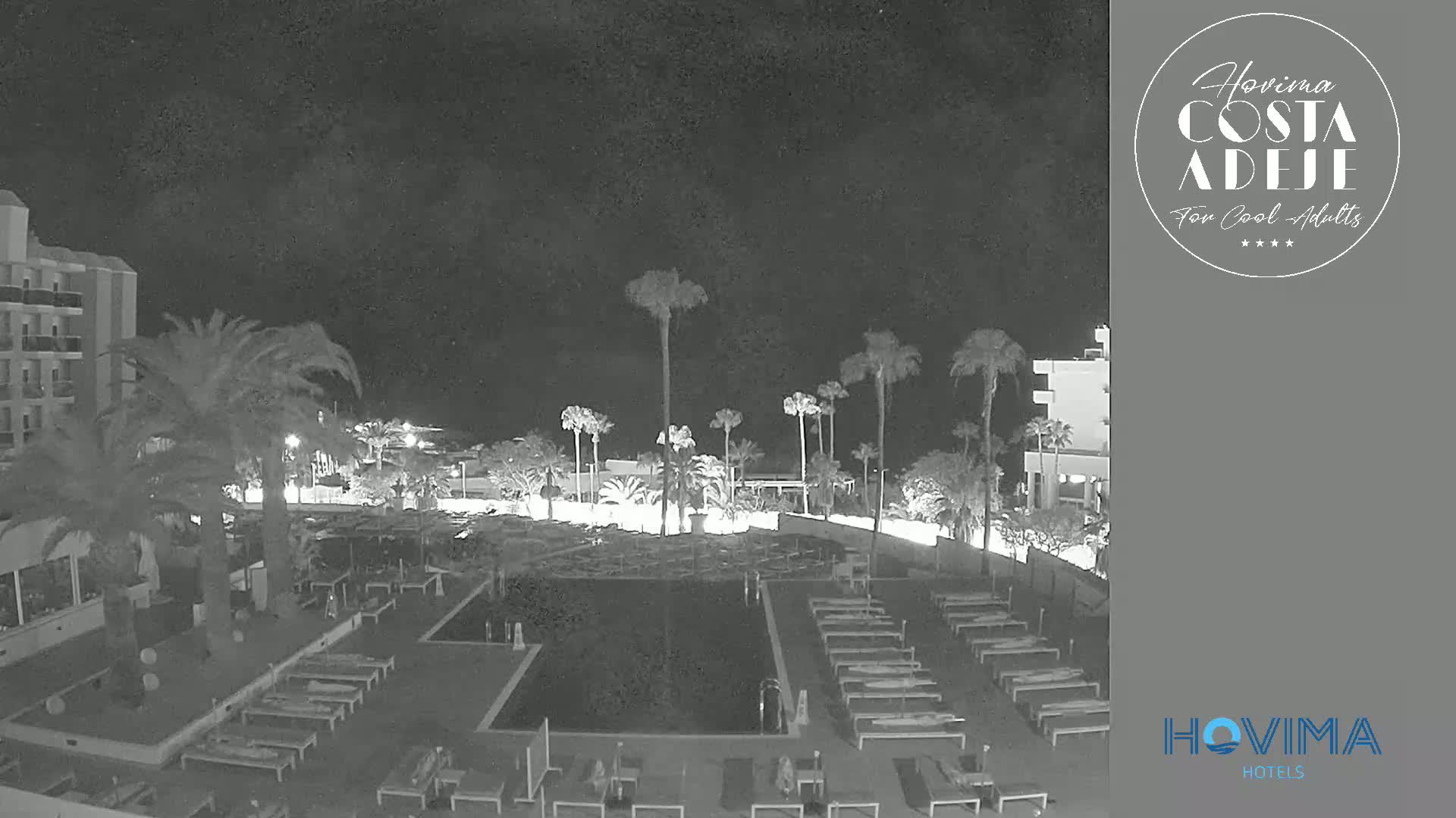 A nighttime, black and white view of a resort pool area with palm trees, lounge chairs, and a building.